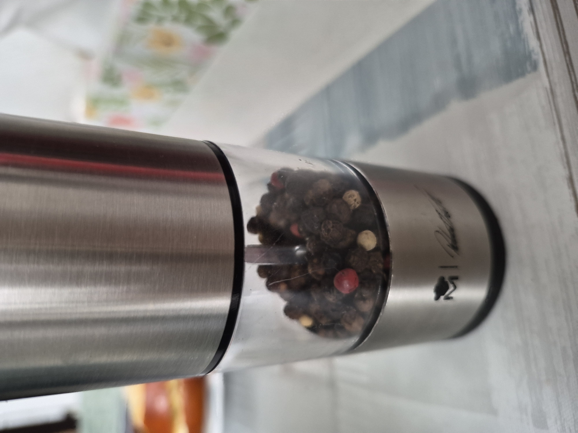 A close-up shot captures a stainless steel and glass pepper grinder filled with peppercorns. The grinder has a metallic top half and a transparent bottom section, allowing the contents to be visible. The peppercorns are predominantly dark, with a few lighter-colored ones, including one distinctly red peppercorn. A logo, an 'M' inside a cap shape, and a signature are visible on the metal base of the grinder. The background is blurred, showing hints of a patterned fabric and a light-colored surface. The image is taken from a slightly elevated angle.