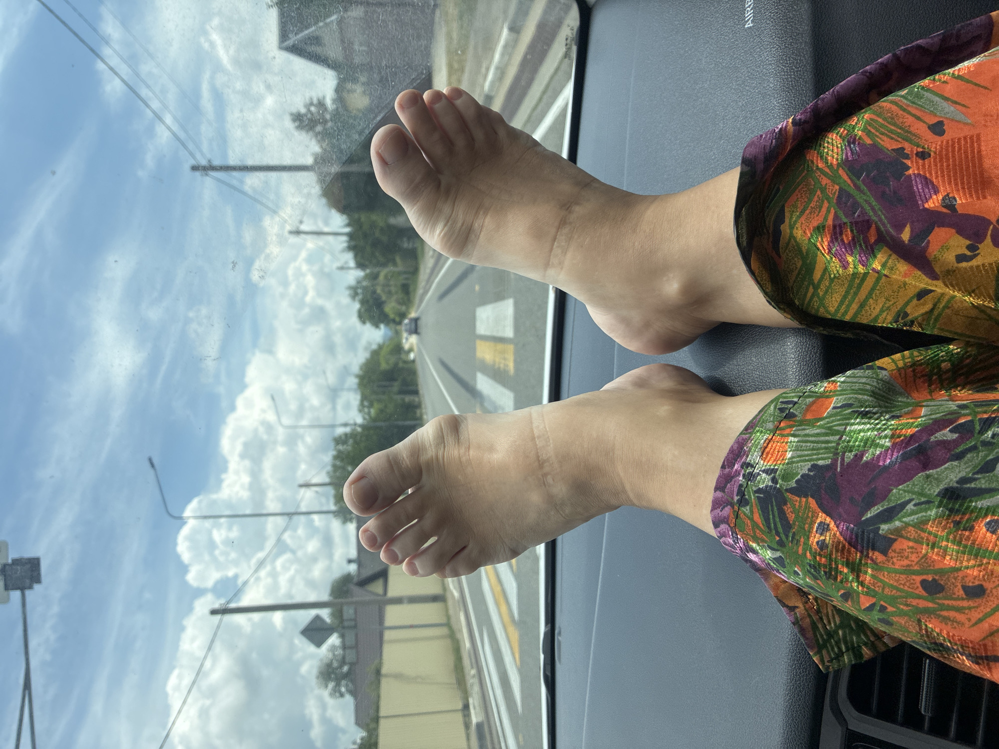 The image shows a first-person view of a person's bare feet resting on the dashboard of a car, with their legs bent. The person is wearing brightly colored patterned pants. The view through the windshield shows a road with a pedestrian crossing, streetlights, power lines, trees, houses, and a cloudy sky. A car is visible in the distance on the road. The location is the city of Tekos, Russia.