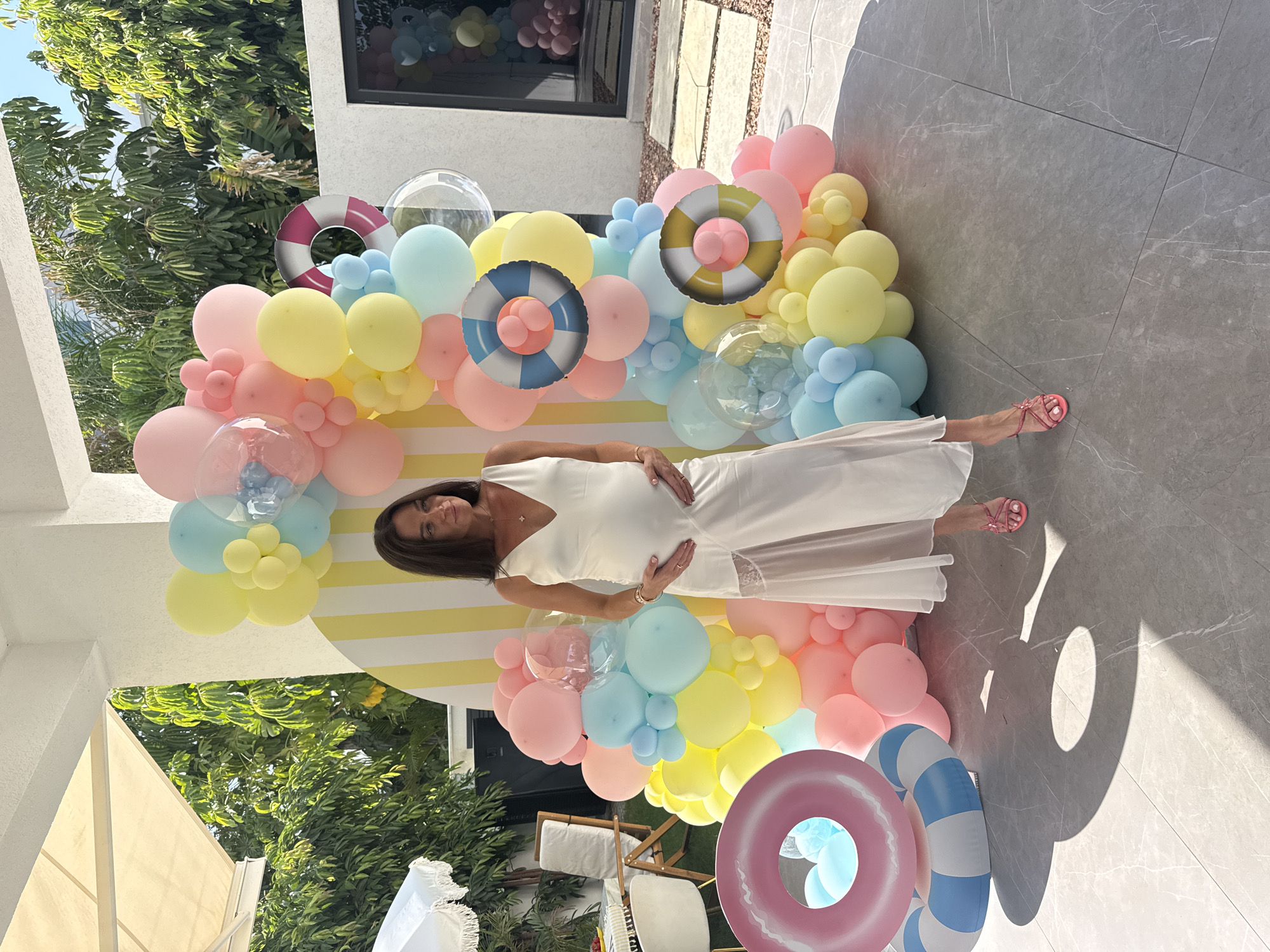 In Agios Tychona Community, Cyprus, a pregnant woman in a white satin dress stands in a sunlit outdoor setting. She is positioned in front of a large balloon archway adorned with pastel-colored balloons in shades of pink, yellow, and blue, along with transparent balloons. Two inflatable rings, one pink and white striped and another with a yellow and white striped pattern, are placed on the ground near the balloon display. The woman is holding her pregnant belly with both hands and is looking away from the camera. Her hair is dark and shoulder-length. She is wearing pink heeled sandals. The background reveals lush green trees and a white umbrella. The ground is paved with gray tiles that have a marble-like pattern. The scene appears to be a celebration or event, possibly a baby shower, given the decorations and the pregnant woman.