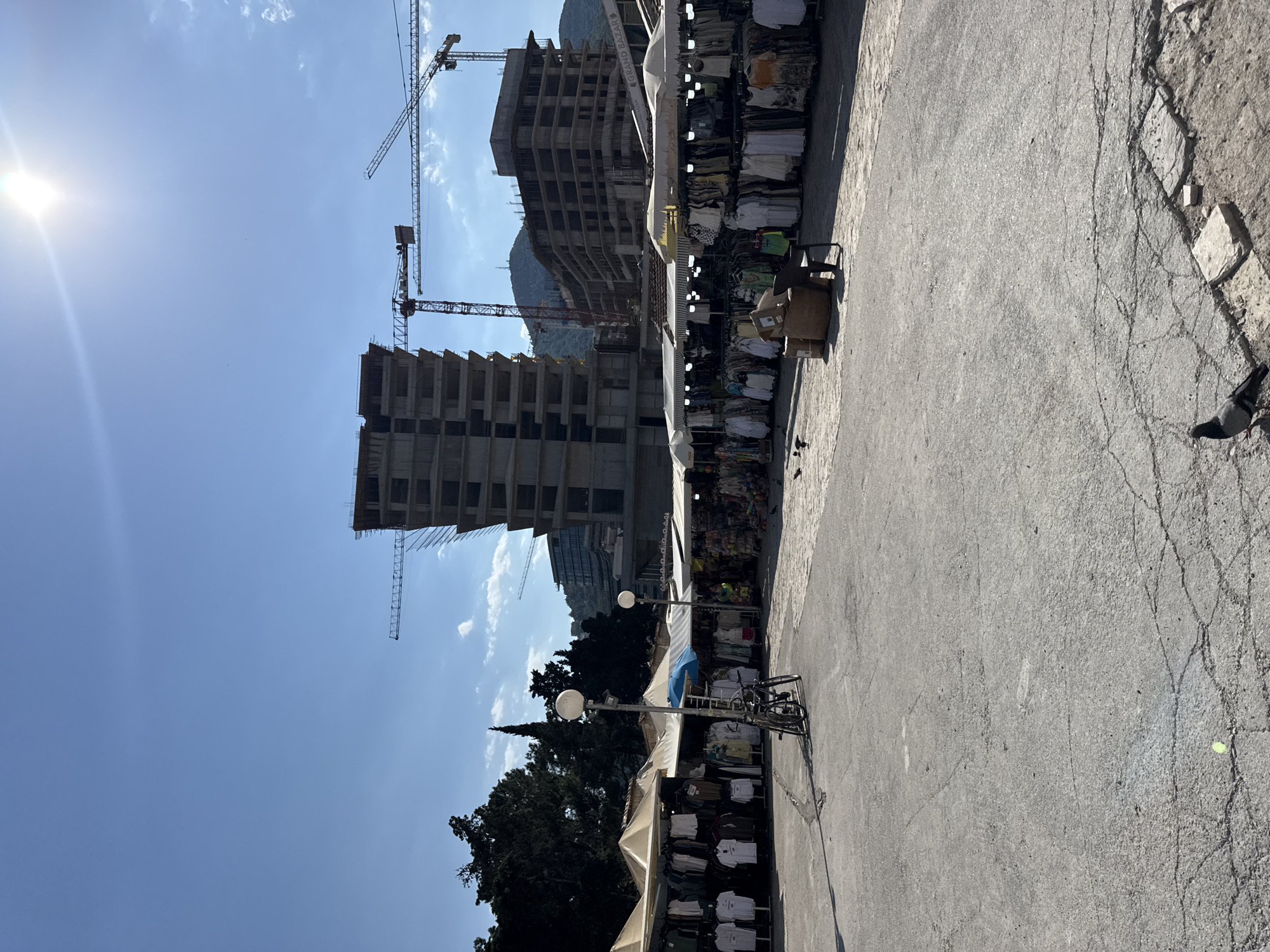 This image depicts an outdoor market scene in Budva, Montenegro, with a construction site in the background. The sun is shining brightly, suggesting it is daytime, and the sky is clear with a few wispy clouds.
In the foreground, there are market stalls displaying a variety of clothing, primarily white t-shirts and other apparel. A bicycle is parked near one of the stalls, and a chair is set up, possibly for a vendor. A person is partially visible in one of the stalls, appearing to be attending to the merchandise. Further down the market, there are some cardboard boxes. A pigeon is seen on the pavement in the lower right corner.
The background is dominated by two large buildings under construction, with cranes extending upwards. These structures suggest ongoing development in the area. A mountain range is visible behind the construction site. Trees are also present, adding some greenery to the scene. The market appears to be set up on a paved area, with some cracked sections in the ground. A street lamp is visible near the market stalls.
The overall atmosphere seems to be one of everyday activity, with the market operating alongside a significant construction project. The bright sunlight and clear sky indicate pleasant weather.