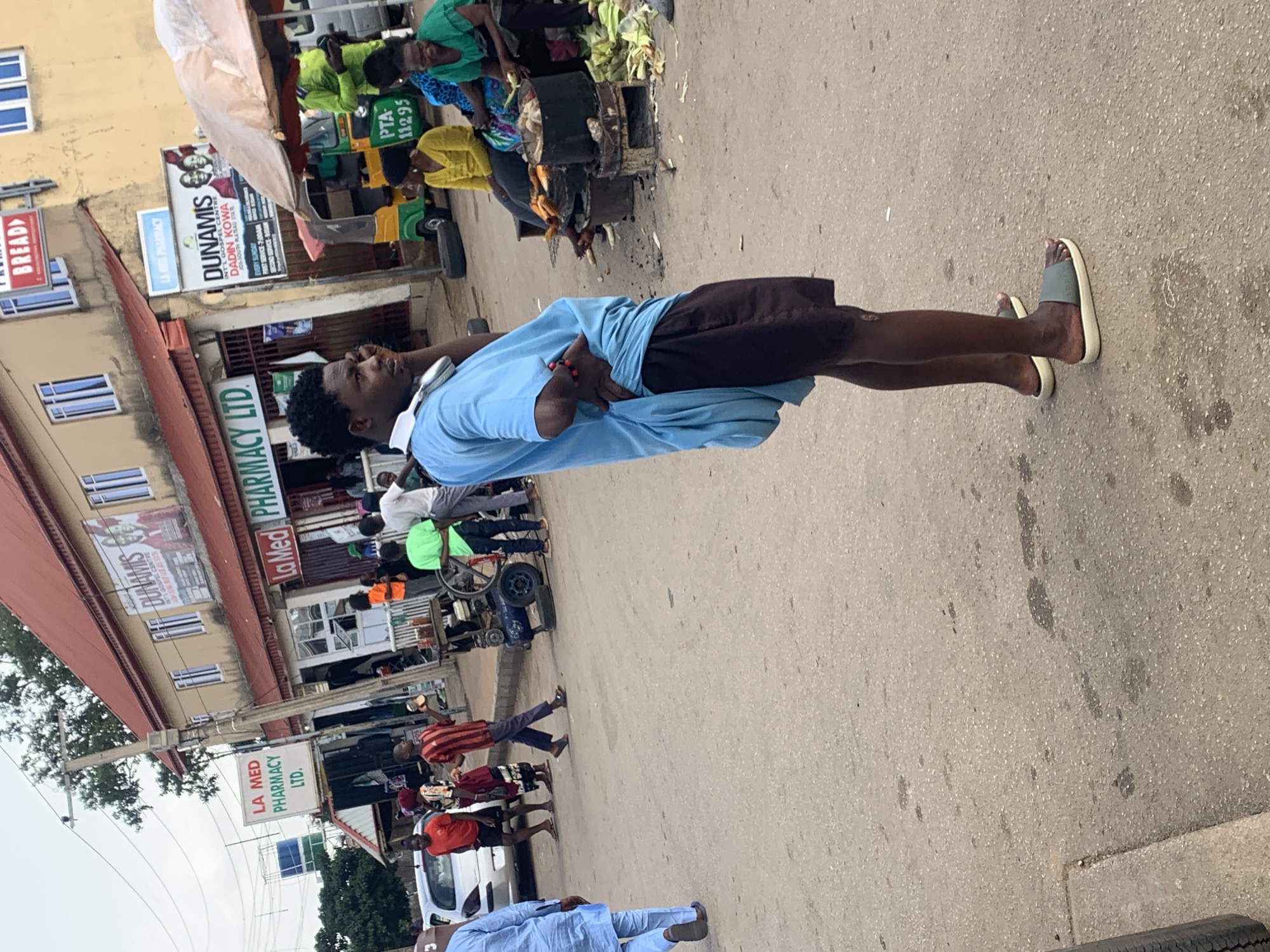 This media file captures a street scene in Gura Top, Nigeria, likely during daytime hours under overcast skies. The focal point is a young man standing in the middle of a paved street, looking to his left. He is wearing a light blue t-shirt and dark shorts, with a pair of white headphones around his neck and a red bracelet on his left wrist. He is also wearing light grey slide sandals.
The background reveals a busy marketplace environment. Several shops line the street, with prominent signage for "LA MED PHARMACY LTD." and "DUNAMIS INT'L GOSPEL CENTRE." Other visible text includes "BREAD" and "PTA 11295." Various individuals are scattered throughout the scene, some appearing to be customers or vendors. To the left, a stall with a large, pale canopy is visible, and further back, people are gathered around what looks like food stalls. A few vehicles, including a white car and a bicycle with a cart attachment, are also present. The overall atmosphere suggests a typical urban street in Nigeria, characterized by commerce and pedestrian activity.