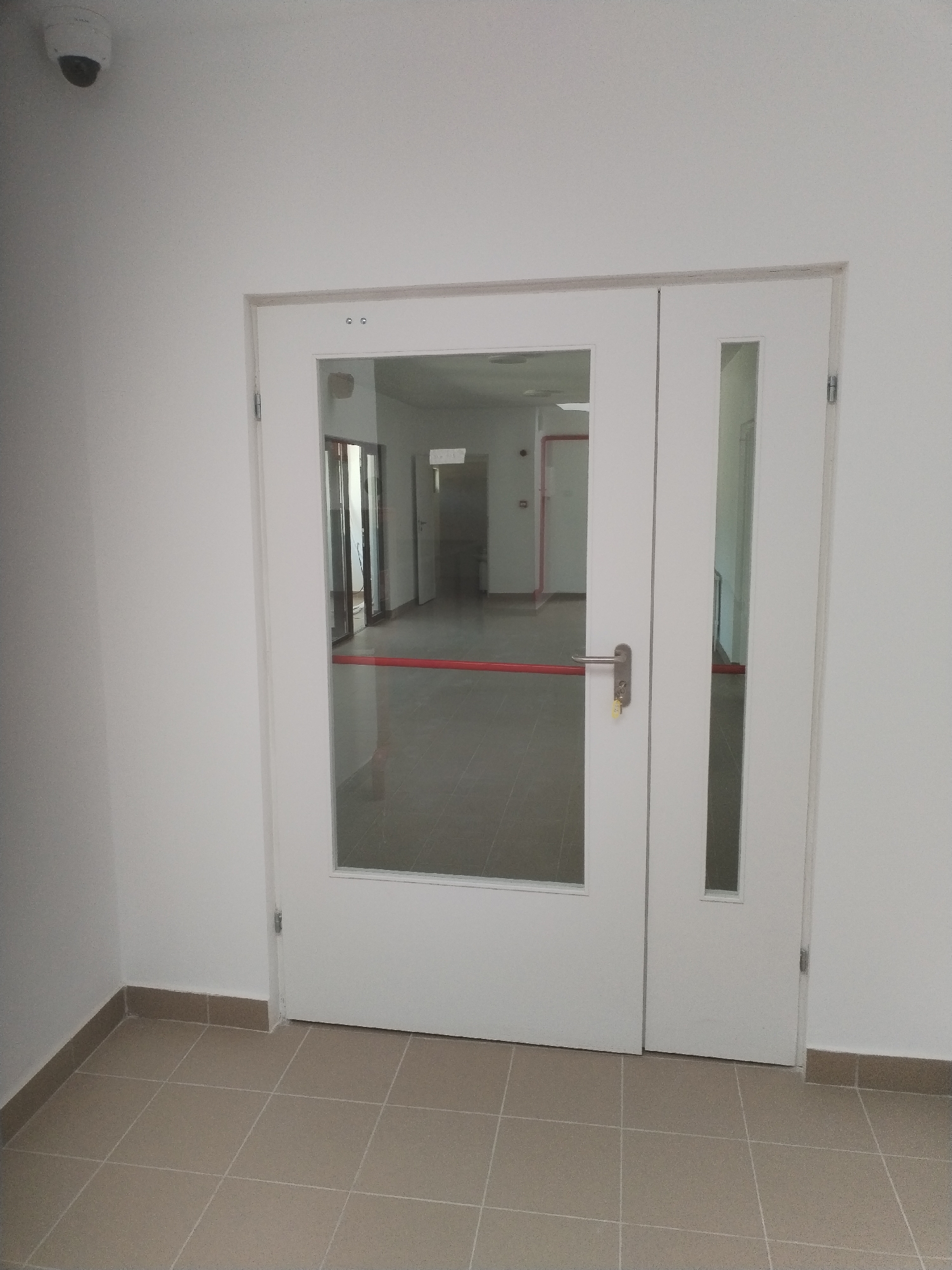The image depicts a hallway with a set of double doors, one of which is partially open. The doors are white and have glass panels. A red bar extends across the larger glass panel, likely a push bar for emergency exits. On the left side of the hallway, near the ceiling, a white security camera is visible. The floor is tiled with square, beige tiles. The walls are painted white. The scene appears to be indoors, in a public or commercial building.