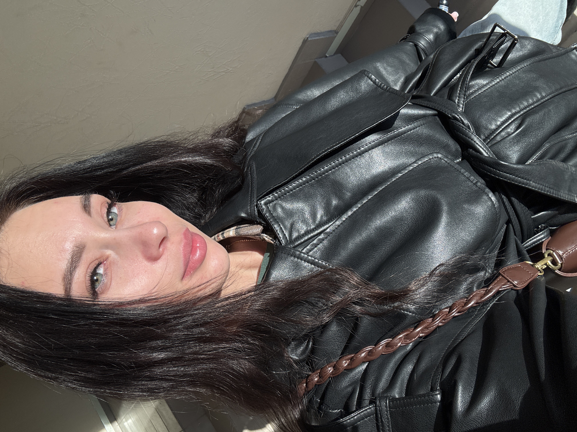 A woman with dark hair and light eyes is the subject of a close-up photograph. She is wearing a black leather jacket with a tie belt, and a brown braided strap is visible across the jacket. The photo appears to be taken outdoors in daylight. The background is a light, neutral color, possibly a wall or ceiling. There is no information provided in the image or prompt regarding a specific location like Chaikovsky, Russia.