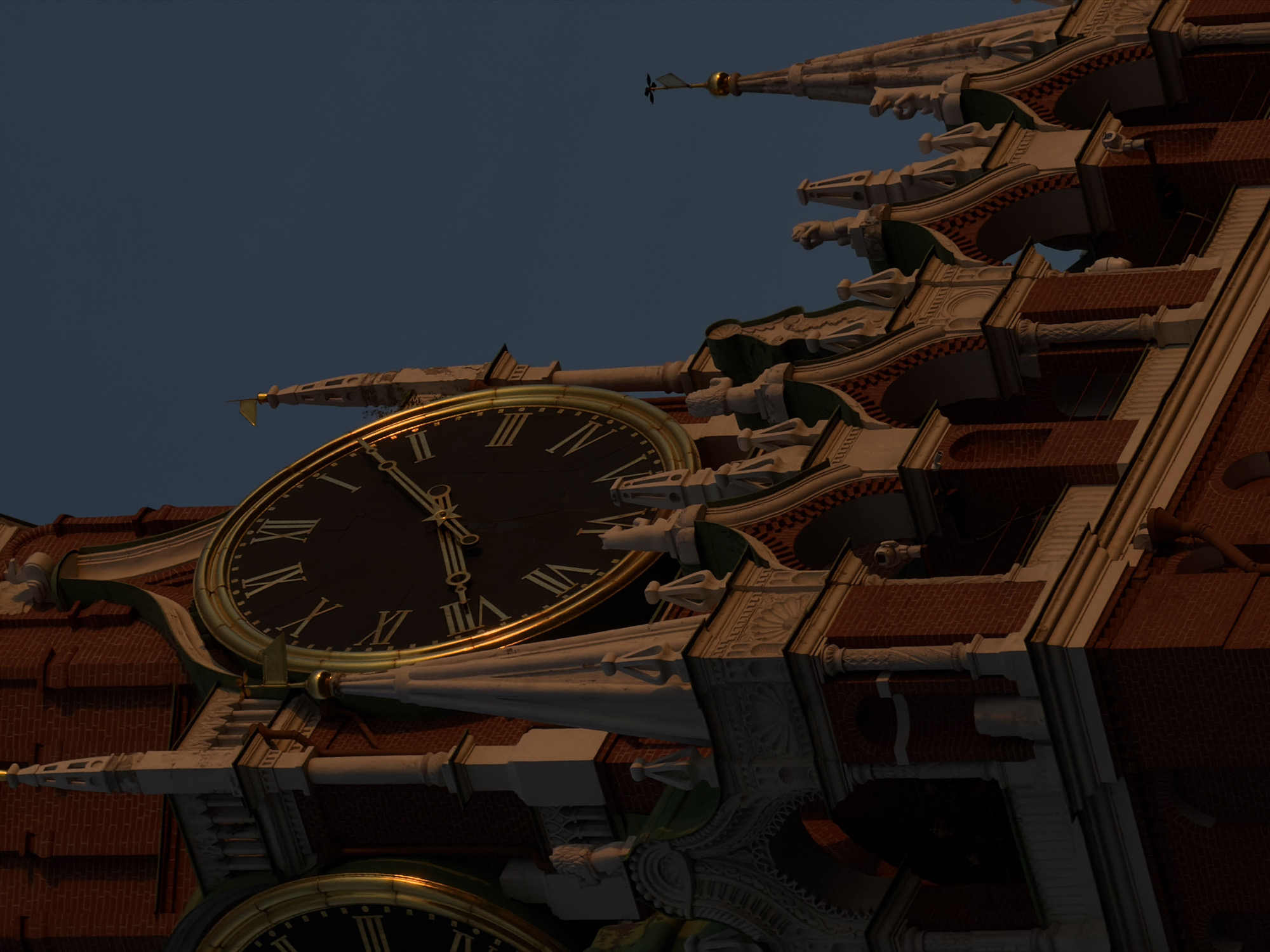 The image depicts a close-up view of the Spasskaya Tower clock located in Moscow, Russia, against a deep blue sky. The tower's architecture features red brickwork and elaborate white stone carvings, including gargoyles. The clock face is large and circular, with Roman numerals and golden trim. The time displayed on the clock face is approximately 10:24. The image suggests either dusk or dawn due to the lighting. The angle suggests a ground-level view looking up at the tower. Another clock face is partially visible in the lower-left corner.