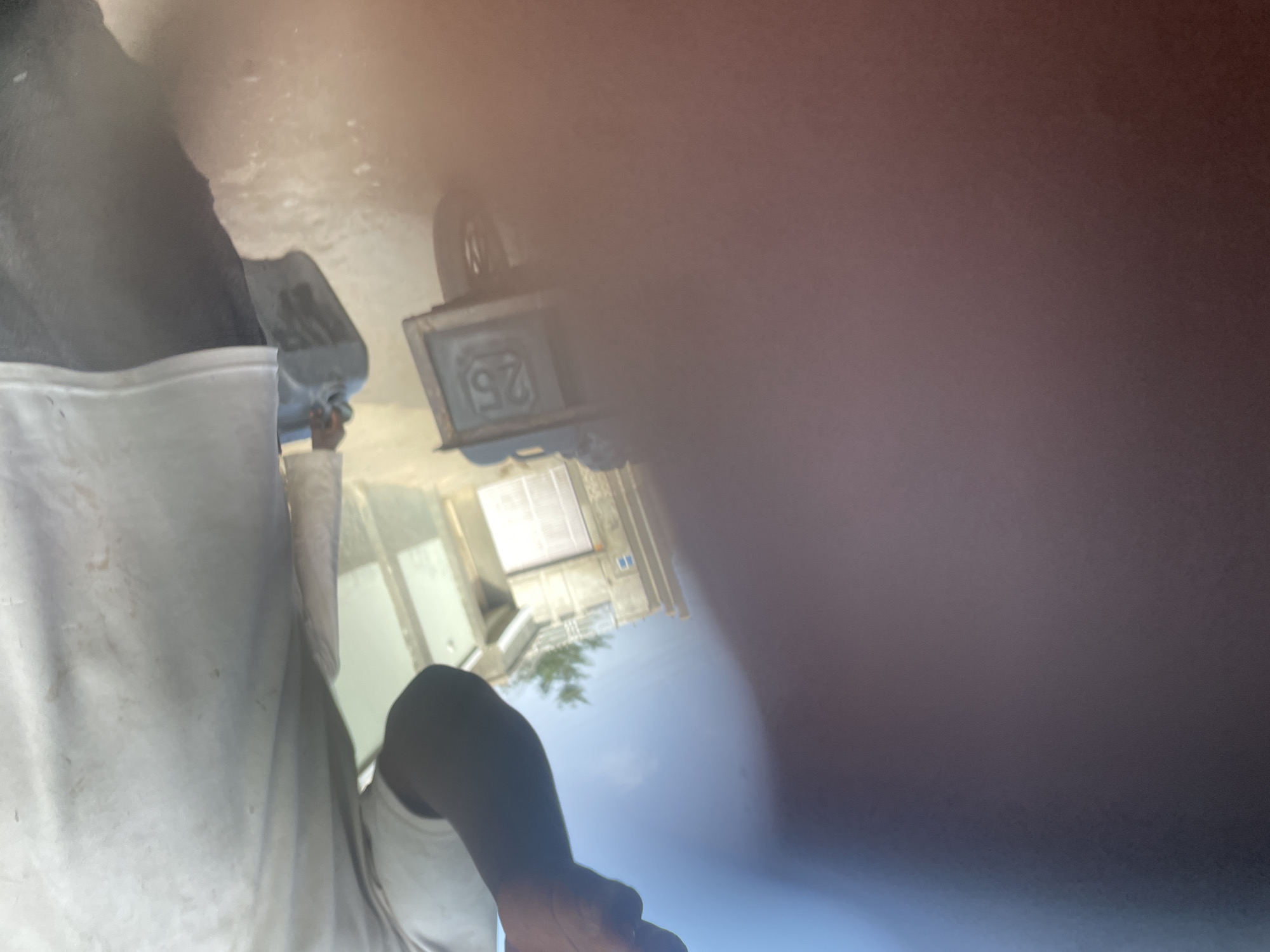 The image depicts an outdoor scene, likely during daytime under a clear sky. A significant portion of the right side of the image is obscured by a large, blurry, reddish-brown object, possibly a finger or part of the camera lens.
In the foreground, on the bottom right, a person is partially visible, wearing a white t-shirt and a dark garment, possibly overalls or a jacket, over it. Their right arm is bent, with the hand visible. The white fabric of their shirt shows some dirt or stains.
Further into the scene, towards the upper left, another person is seen holding a blue plastic container or jug. They appear to be engaged in an activity involving this container, possibly pouring or carrying its contents.
In the midground, towards the upper right, there is a dark, box-like object with the number "25" prominently displayed in white. Above this object, a tire or wheel is partially visible. The background features several buildings with windows, suggesting an urban or industrial setting, along with some green foliage from a tree, set against a bright blue sky. The lighting indicates it is a sunny day.