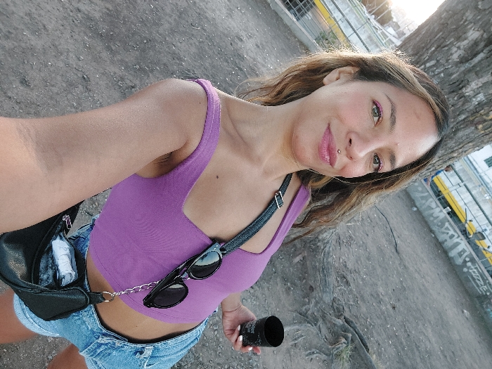 A female subject, appearing to be in her 20s or 30s, is depicted in a selfie taken outdoors in an urban park in Buenos Aires, Argentina. She has a light-brown complexion, long wavy brown hair with lighter streaks, and light-colored eyes. Her face displays a slight smile, with pink lipstick and pink eyeliner visible. A silver labret piercing is present above her upper lip. She is wearing a purple sleeveless crop top and blue denim shorts. Black sunglasses are clipped to the strap of her top, connected by a silver chain to a small black shoulder bag worn across her body. In her right hand, she holds a black cup adorned with white designs; her fingernails are painted white. The immediate background shows a large tree trunk to the left and exposed roots on the dirt ground. Further in the background, a yellow structure is visible above a concrete wall covered in graffiti, including the word "PIZZA." A light-colored building is partially visible beyond the graffiti-covered wall. The lighting suggests late afternoon or early evening.