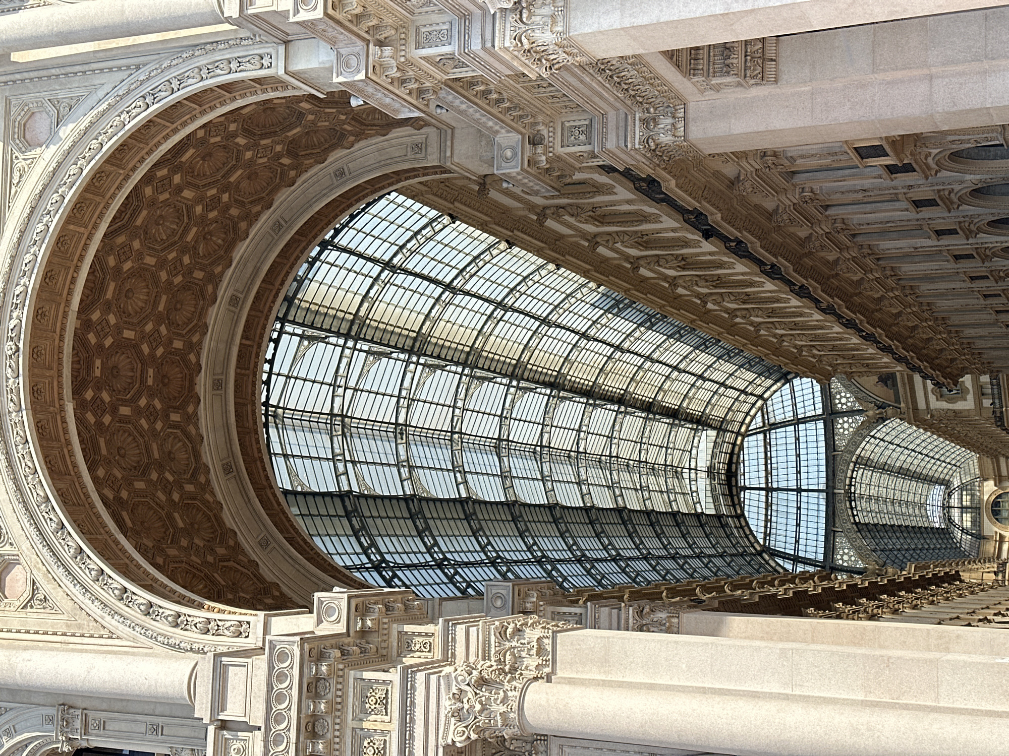 The image depicts the interior of the Galleria Vittorio Emanuele II in Milan, Italy. The perspective is looking upwards towards the grand glass and iron dome and ceiling. Sunlight streams through the vast expanse of glass panels, illuminating the intricate architectural details. Ornate stucco work and decorative moldings adorn the arched structural elements and the facades of the shops lining the gallery. The scene is devoid of people, focusing solely on the majestic architecture and the interplay of light and shadow within the iconic Milanese landmark. The detailed carvings and the repetition of architectural motifs are prominent features.