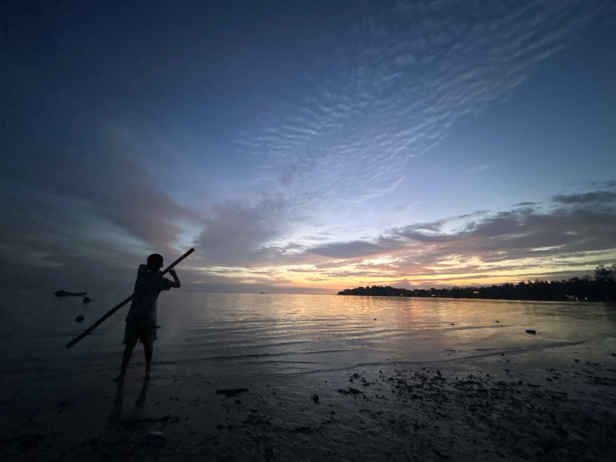 A silhouette of a person stands on a beach at dusk in Ko Pha-ngan District, Thailand, holding a long stick at an upward angle. The sky is a gradient of deep blue at the top, transitioning to lighter blues and oranges near the horizon, with scattered clouds. The calm sea reflects the colors of the sky. The beach is dark and sandy, with some scattered debris. A dark, tree-lined landmass is visible in the distance.