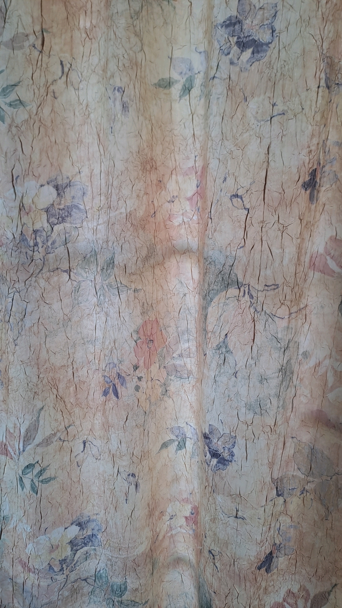 The image is a close-up of a textured, creased fabric with a faded floral pattern. The colors are muted, with shades of beige, pale pink, light green, and dark blue. The fabric appears to be hanging, as indicated by vertical folds and shadows. There are no people or specific objects visible beyond the fabric itself. The image does not contain any information that would allow for identification of its location in Kolomyia, Ukraine.
