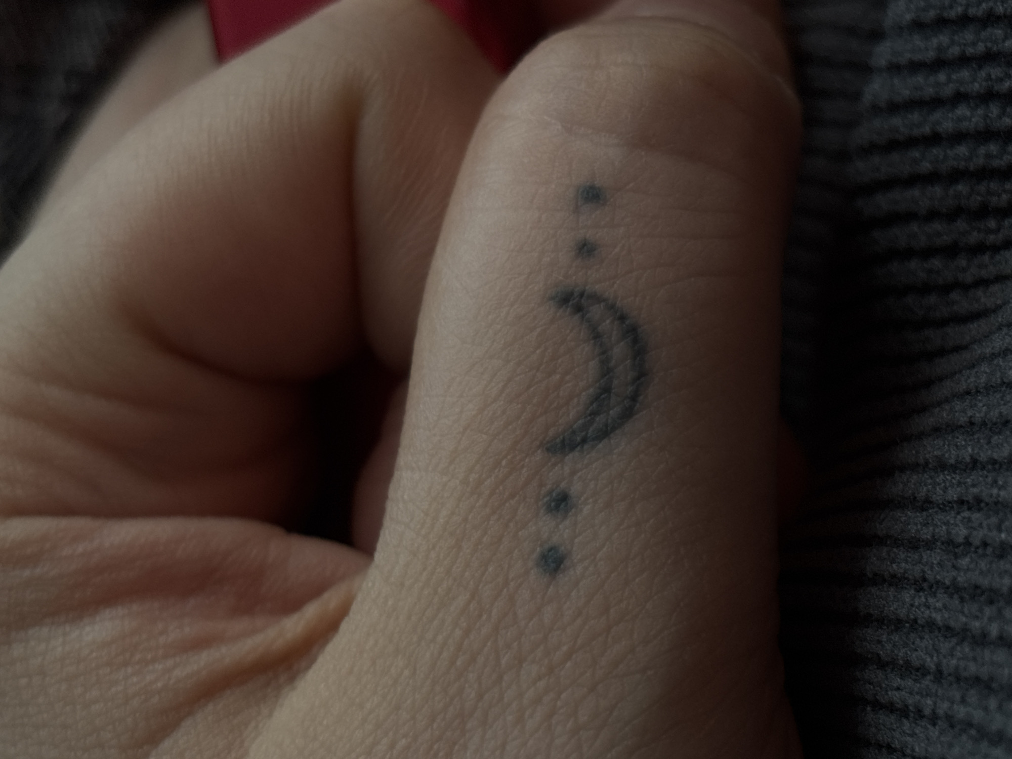 This media file is a close-up photograph of a person's finger, specifically the side of the thumb, showing a small, minimalist tattoo. The tattoo depicts a crescent moon with three dots arranged vertically above it. The skin texture is visible, showing fine lines and pores. The lighting suggests an indoor setting, possibly with natural light from a window given the soft illumination. The background is blurred, with hints of a dark, ribbed fabric, possibly clothing or a blanket, and a bright red object out of focus in the upper left corner. The image is focused on the tattoo and the immediate skin area, with the rest of the hand and background out of focus. No text is visible in the image. The overall impression is intimate and personal, highlighting the detail of the tattoo on the skin.