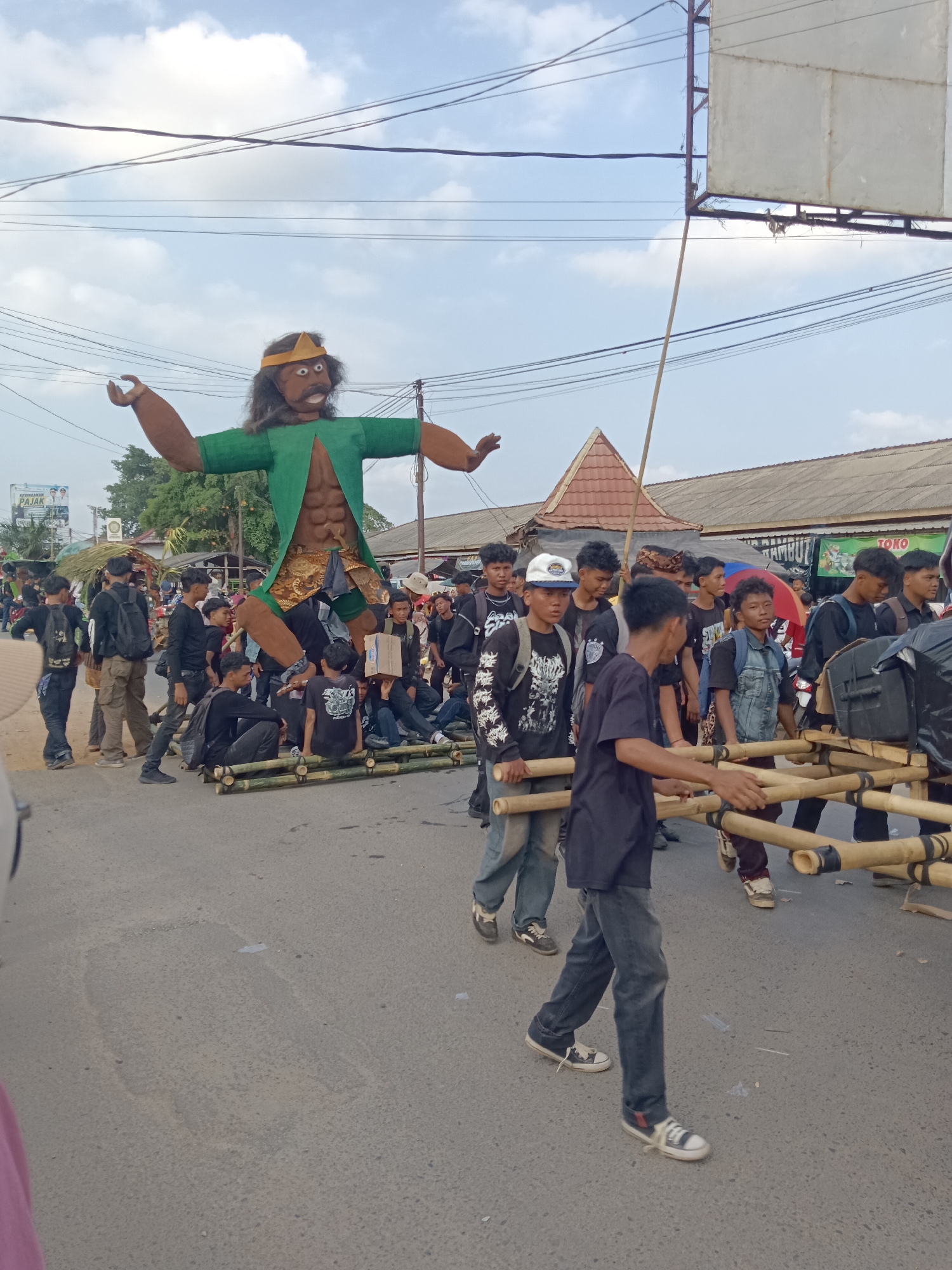 In Tegal Gondo, Indonesia, a procession is underway on a paved street under a cloudy sky. The central focus is a large effigy of a human figure, approximately ten feet tall, dressed in a green vest and loincloth, with a golden headdress. The effigy is supported by a bamboo structure carried by a group of young men. These men are dressed in casual attire, predominantly black t-shirts and jeans, and are walking in formation, some carrying the bamboo poles that bear the effigy. Other individuals, also dressed casually, are visible in the background, some walking alongside the procession and others gathered on the sides of the street. Buildings with corrugated roofs and awnings line the street, suggesting a commercial or residential area. Overhead, power lines crisscross the sky. The scene captures a cultural event or celebration taking place in a community setting.