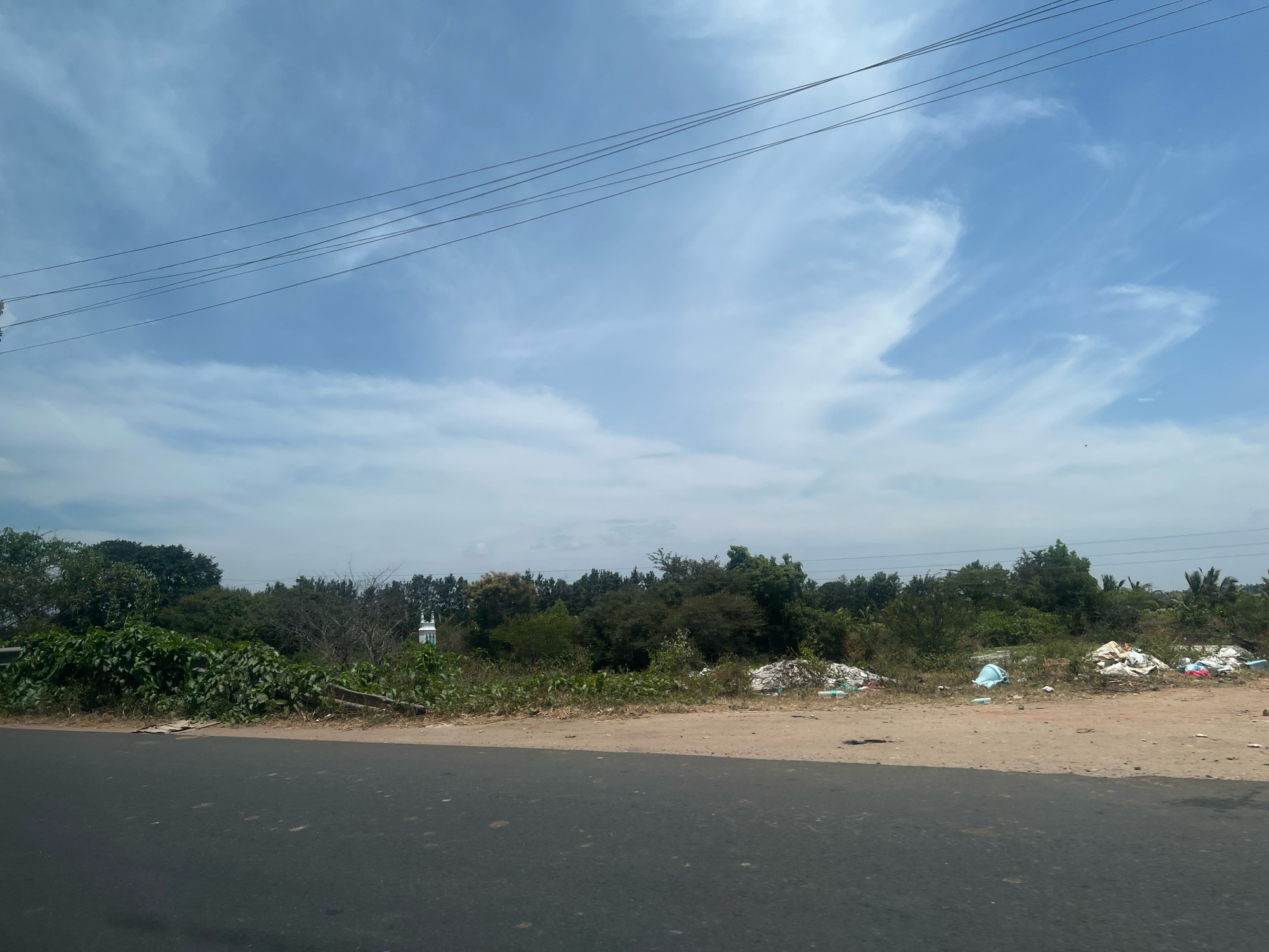 This image depicts a roadside scene in Kampalapura, India, characterized by a paved road in the foreground and a natural environment beyond. The road appears to be a two-lane asphalt surface with a dirt shoulder. The shoulder is littered with various debris, including plastic bags, wrappers, and what appear to be discarded containers.
Beyond the shoulder, there is a dense collection of trees and vegetation, forming a natural barrier. Interspersed within this greenery are signs of human activity. A small white structure with a green accent, possibly a minaret or a part of a religious building, is visible amidst the trees on the left side of the frame. Further to the right, more piles of trash are visible, suggesting a lack of proper waste disposal in the area.
Overhead, a series of power lines are stretched across the bright blue sky, which is dotted with wispy white clouds. The overall impression is one of a rural or semi-rural roadside environment in India, marked by both natural beauty and the presence of litter and infrastructure. There are no people or animals visible in the image. The lighting suggests it is daytime, likely late morning or early afternoon, given the clear sky and shadow patterns.