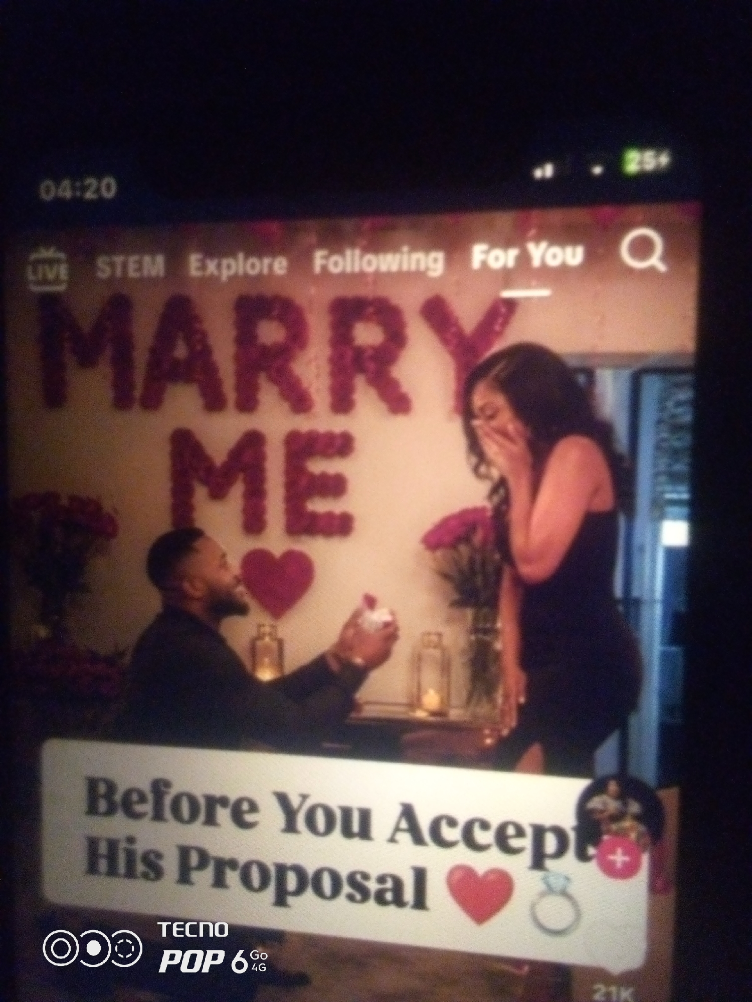 The image shows a screen displaying a video or image with a romantic proposal theme. In the background, the words "MARRY ME" are formed by red heart-shaped objects on a wall. In the foreground, a man is seen presenting a ring to a woman, who appears surprised and is covering her mouth with her hand. Below them, a sign reads "Before You Accept His Proposal". The Tecno Pop 6 4G logo is visible in the bottom left corner, along with a timestamp of 04:20 in the top left and a network indicator of "25+" in the top right. The scene is indoors, with decorative elements like flowers and candles suggesting a celebratory or intimate setting. There is no information in the image to suggest a location in Howayi, Nigeria.