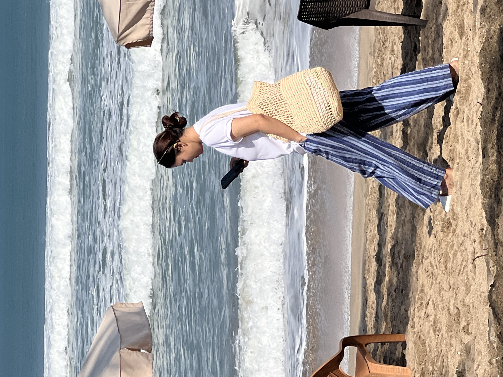 A woman, wearing a white t-shirt and blue-striped wide-leg pants, walks along a sandy beach in Kuta, Indonesia. She is holding a dark-colored smartphone in her left hand and has a woven straw bag slung over her right shoulder. The ocean with gentle waves is visible behind her, and two light-colored beach umbrellas are partially in frame on the left and right sides of the image. A brown plastic chair is situated on the left side of the beach, and a dark chair is visible on the right. The sand is light brown with some scattered debris. The sky is a clear blue.
