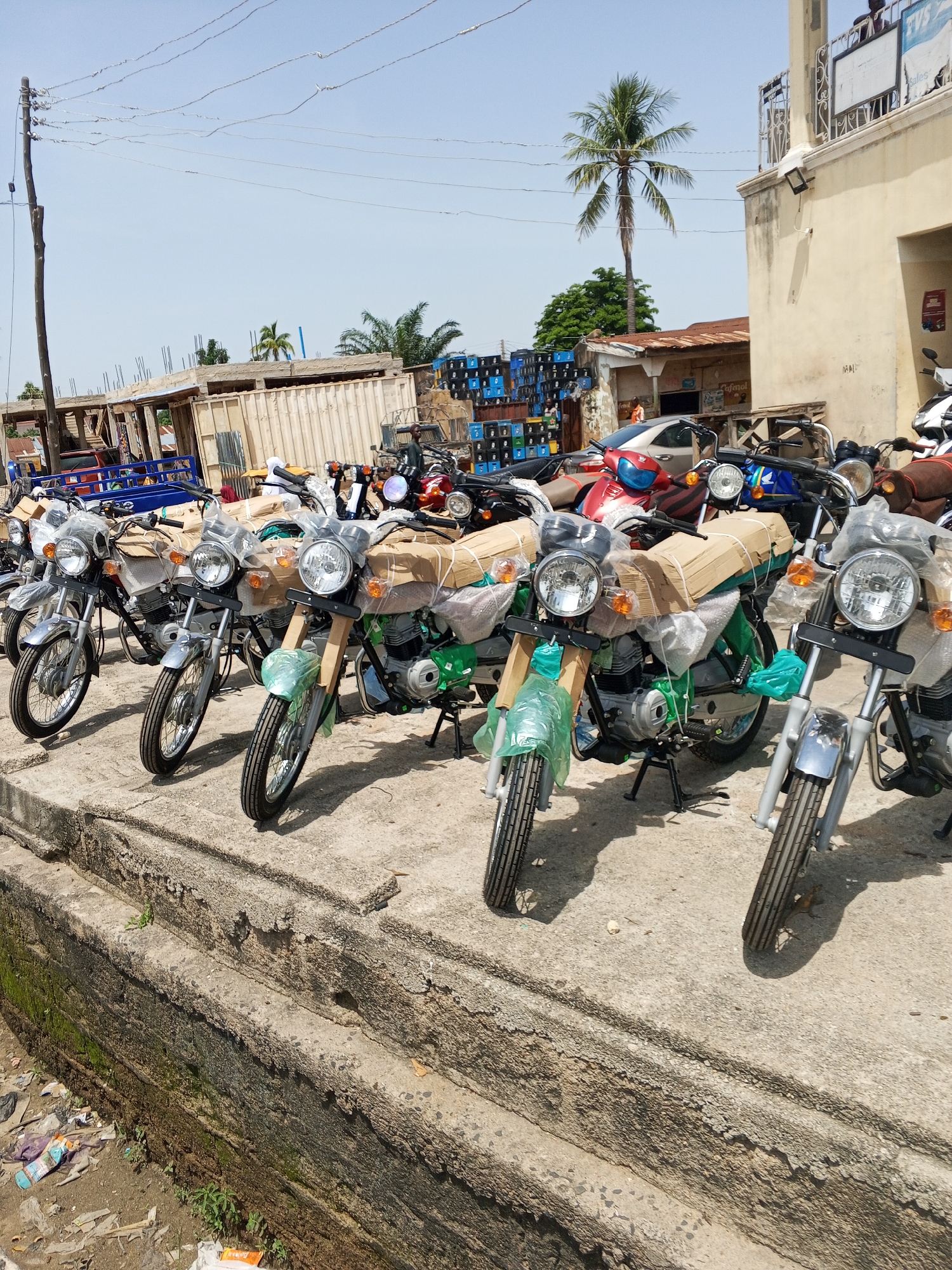 In Jalingo, Nigeria, a row of new motorcycles are parked on a concrete ledge next to a building with a pale yellow facade. The motorcycles are predominantly green and silver, with many covered in clear plastic packaging and brown cardboard. Some motorcycles have their kickstands down, while others appear to be leaning. In the background, stacks of black crates are visible, along with a partial view of a construction site and palm trees. The ground in front of the ledge is littered with debris, including plastic bottles and wrappers. A power pole with wires stretches across the top of the frame.
