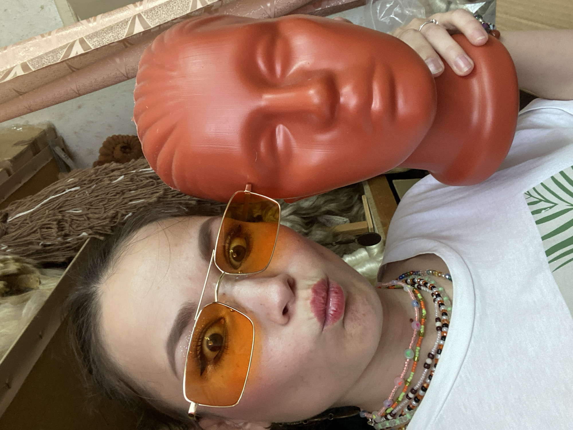 In a dimly lit room, a woman with dark hair pulled back is taking a selfie, holding a red mannequin head. She is wearing large, orange-tinted sunglasses, a white t-shirt, and multiple colorful beaded necklaces. Her face is close to the camera, with her lips pursed and her eyes wide, reflecting the bright lights. The mannequin head is positioned next to her face, partially obscuring it. In the background, there are shelves with various objects, including what appears to be a furry costume or wig and rolls of fabric or wallpaper. The overall impression is a casual, indoor setting, possibly a workshop or a storage area, with no discernible connection to the city of Chaikovsky, Russia.