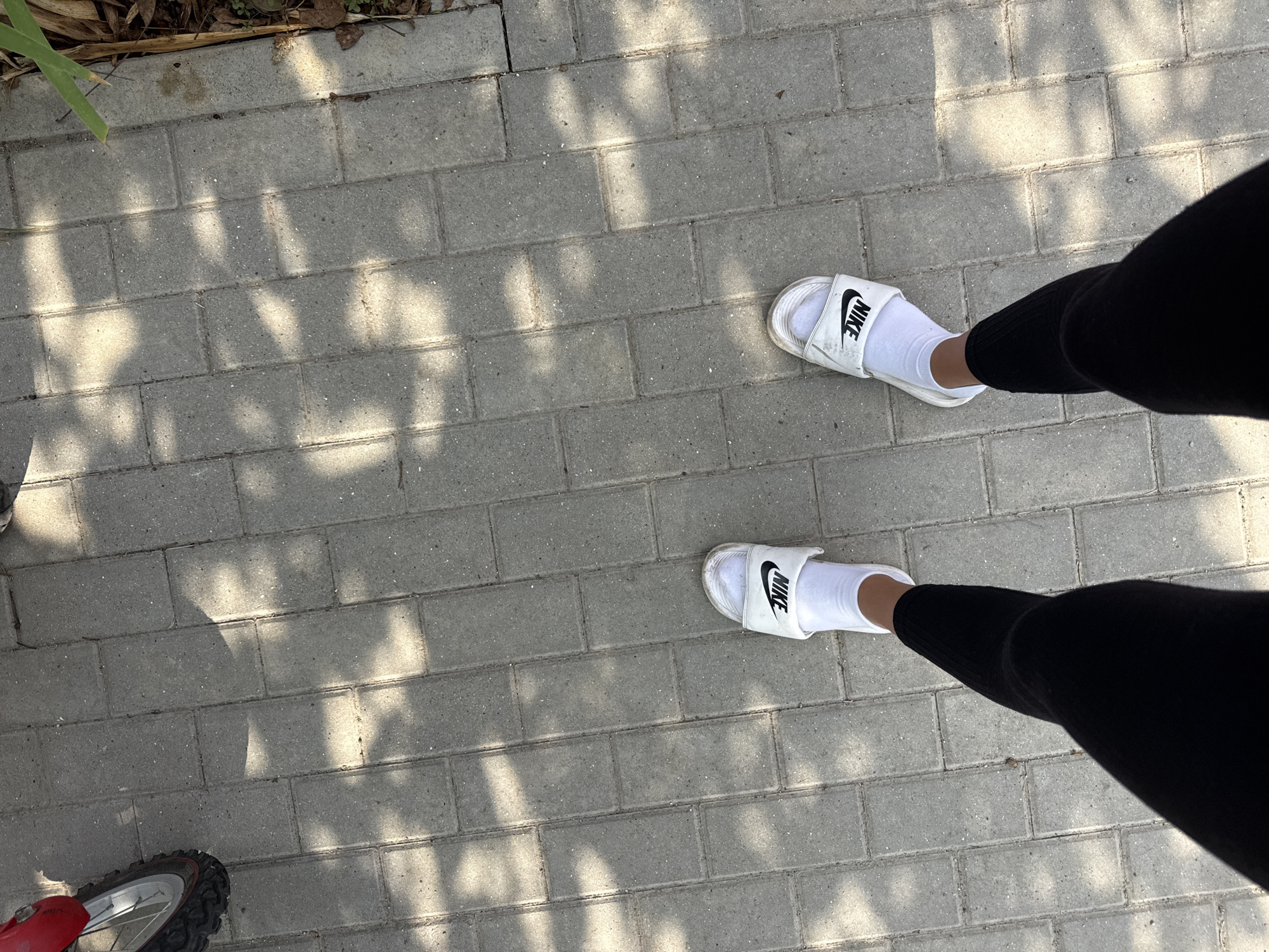 A first-person perspective shot shows a person wearing black leggings, white socks, and white Nike slides standing on a gray brick sidewalk. The sidewalk is partially covered in dappled sunlight and shadow from overhead foliage. In the upper left corner of the image, a portion of a red bicycle wheel is visible. The scene is set outdoors. The location is Basarabeasca, Moldova.