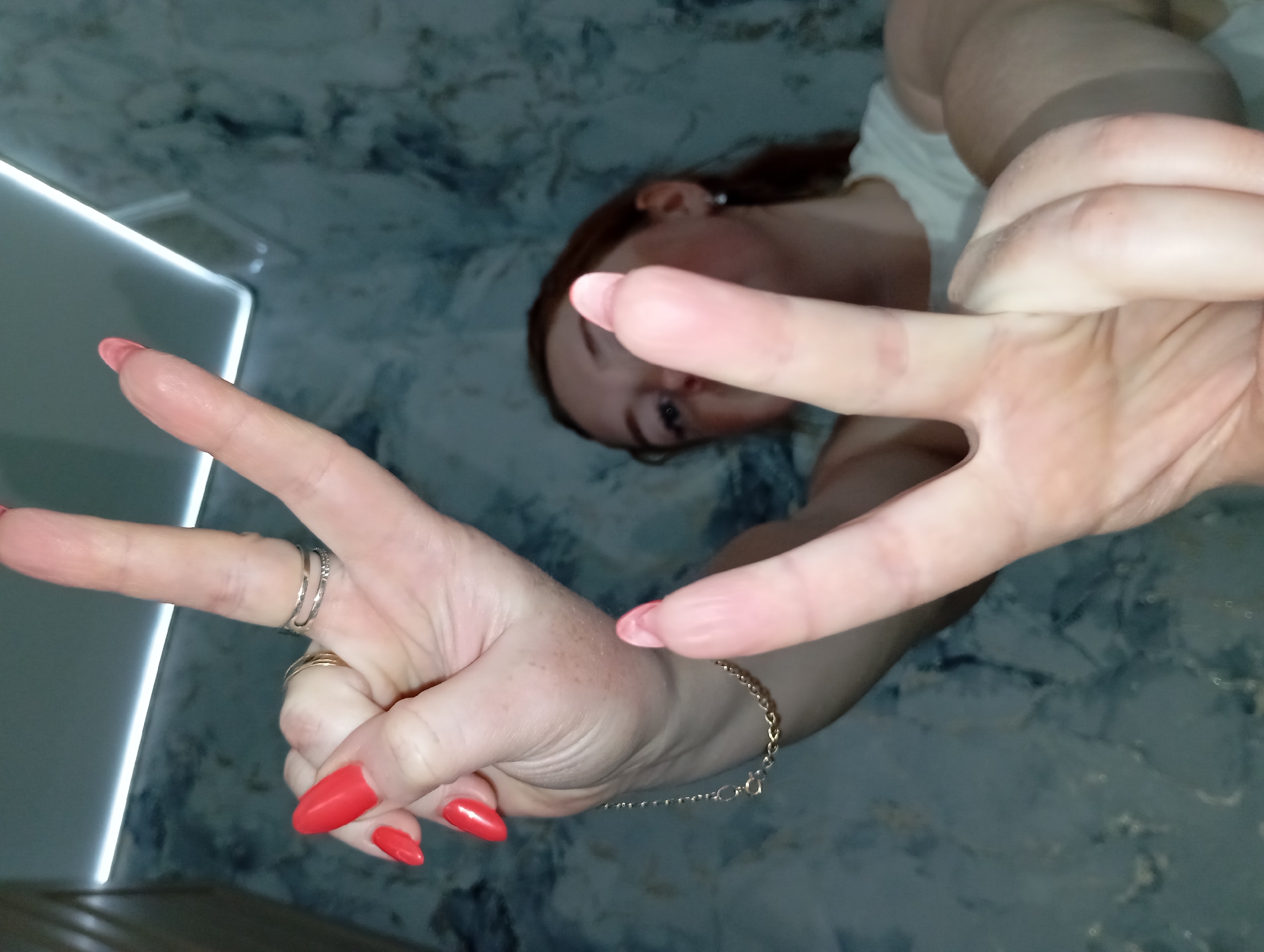 A fair-skinned person with reddish-brown hair, wearing a white top and possibly glasses, is lying on a marbled surface. The person is holding up two hands in peace signs. The hands are adorned with rings on the middle finger of the left hand and a gold bracelet on the wrist of the right hand. The fingernails are painted red. A corner of a light-colored, possibly metallic object is visible in the lower left corner. The location is unknown.