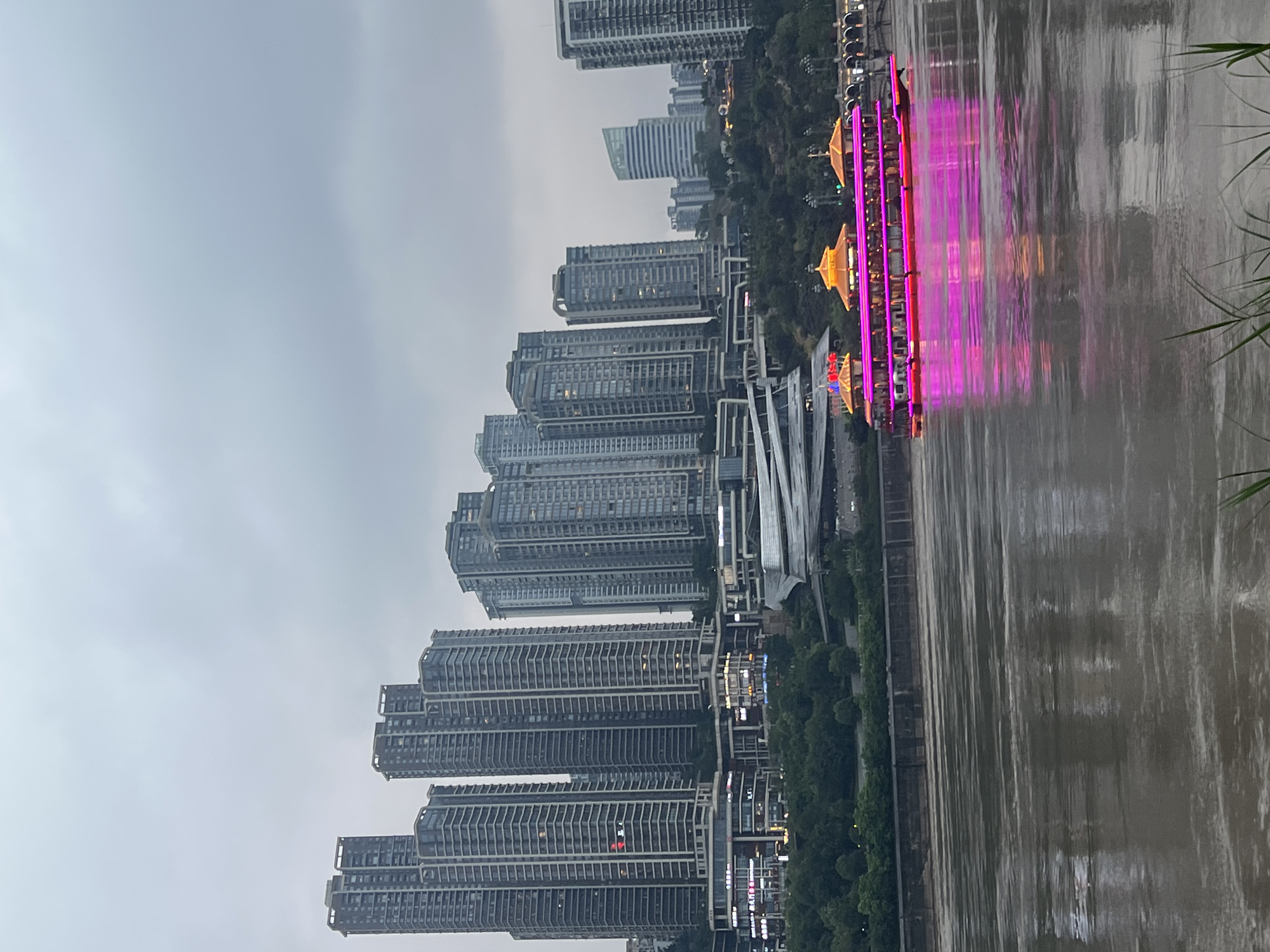 This image shows a view of the Nan'an District in China, featuring a skyline of modern skyscrapers along a waterfront. A brightly lit, multi-decked boat with prominent pink and yellow lights is on the water in the foreground, its reflection shimmering on the surface. The water appears slightly murky, suggesting it is a river. Behind the boat and along the shoreline, there are green trees and various structures, including what appears to be a large, modern building with a distinctive roofline. The sky is overcast with a dull, gray hue. The overall scene depicts an urban waterfront at dusk or on a cloudy day, with a focus on the illuminated boat as a prominent element.