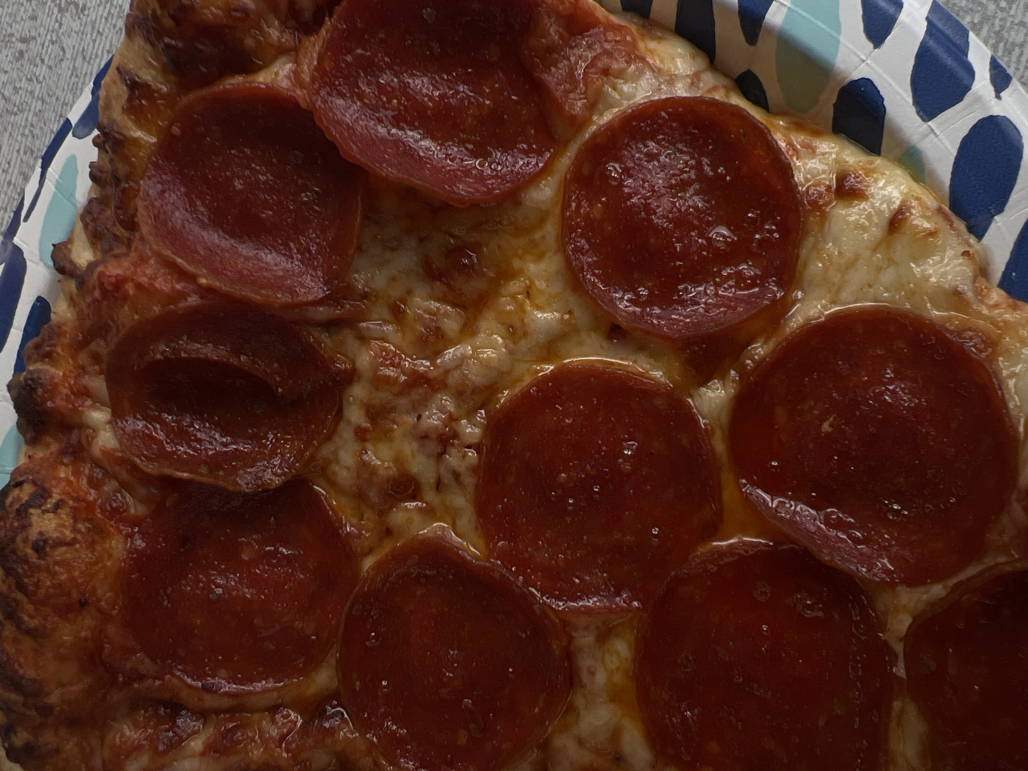 A close-up overhead shot depicts a portion of a pepperoni pizza, served on a disposable paper plate with a blue and white wave pattern. The pizza crust is visible on the left, showing a golden-brown color with varied textures. The cheese is melted, with a slight golden sheen. The pepperoni slices are uniformly circular, red-brown in color, and glistening. The pizza occupies most of the frame. The scene does not suggest any specific location within Mexico City, Mexico. There is no evidence of people or actions.