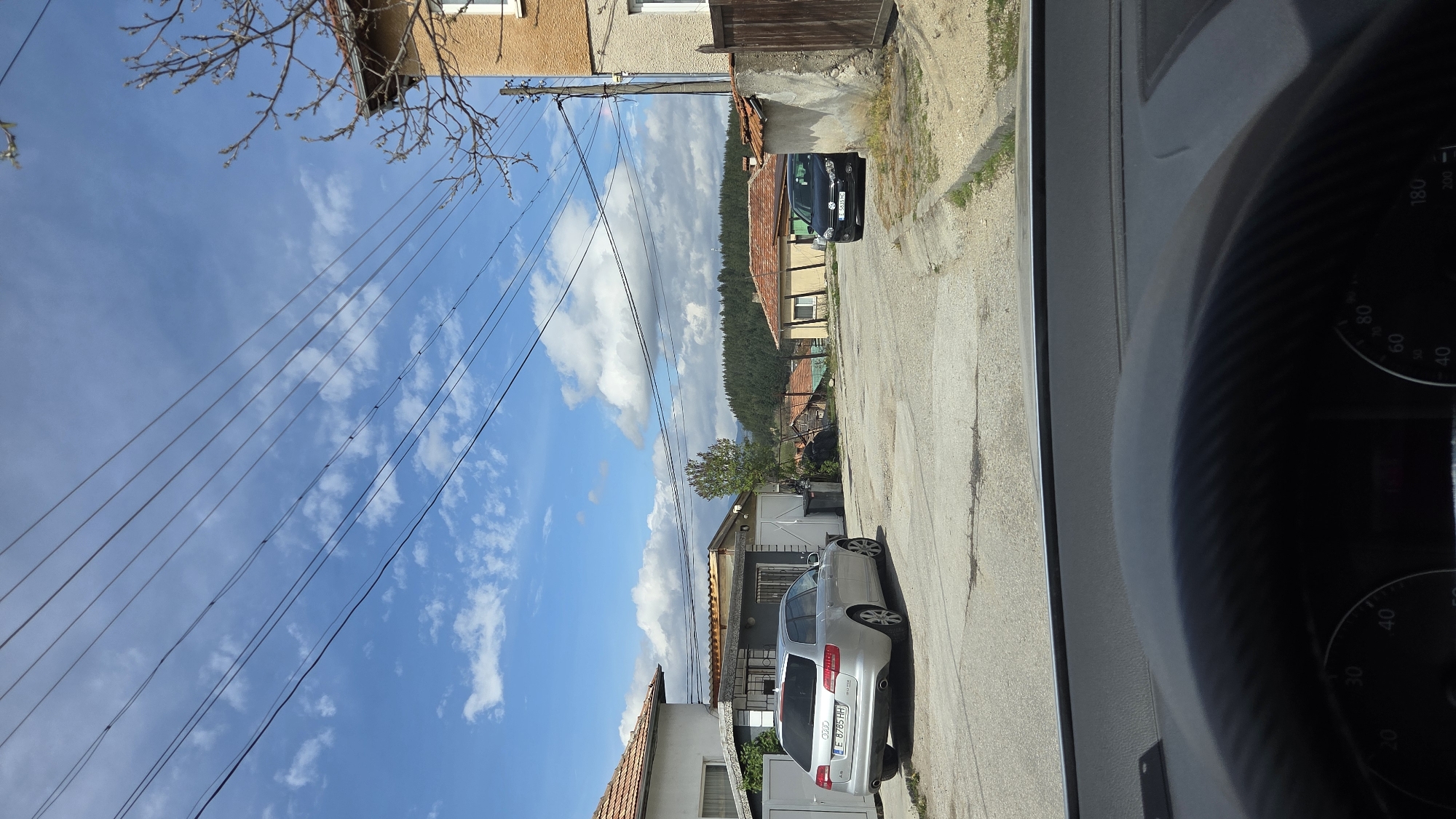 The media file captures a residential street scene, appearing rotated 90 degrees clockwise. The image is taken from inside a vehicle, with a portion of the dashboard visible on the right.
The street, which runs vertically in the rotated image, is lined with houses featuring traditional tiled roofs. On the left side of the street, multiple buildings are visible, including one with a lighter facade in the foreground and older, earth-toned structures further back. Trees are scattered throughout the scene; a bare-branched tree extends into the upper left, and a leafy tree stands further down the street. Numerous power lines are stretched across the bright blue sky, which features scattered white cumulus clouds.
Two cars are parked on the street. A silver Audi station wagon (likely an A4 Avant) is prominent in the mid-ground, displaying a Bulgarian license plate "E 8785 HH", indicating registration in the Gabrovo region. Further down the street, a dark-colored hatchback, possibly a VW Golf, is partially visible, showing a Bulgarian "CB" license plate prefix, suggesting Sofia City registration.
The foreground on the right side reveals part of a car's dashboard, including sections of the instrument cluster with speed indicators such as "40", "60", "80", "180", and "200".
The scene depicts a sunny daytime with fair weather in what appears to be a residential area, likely a town or village in Bulgaria. No people or specific activities are visible.