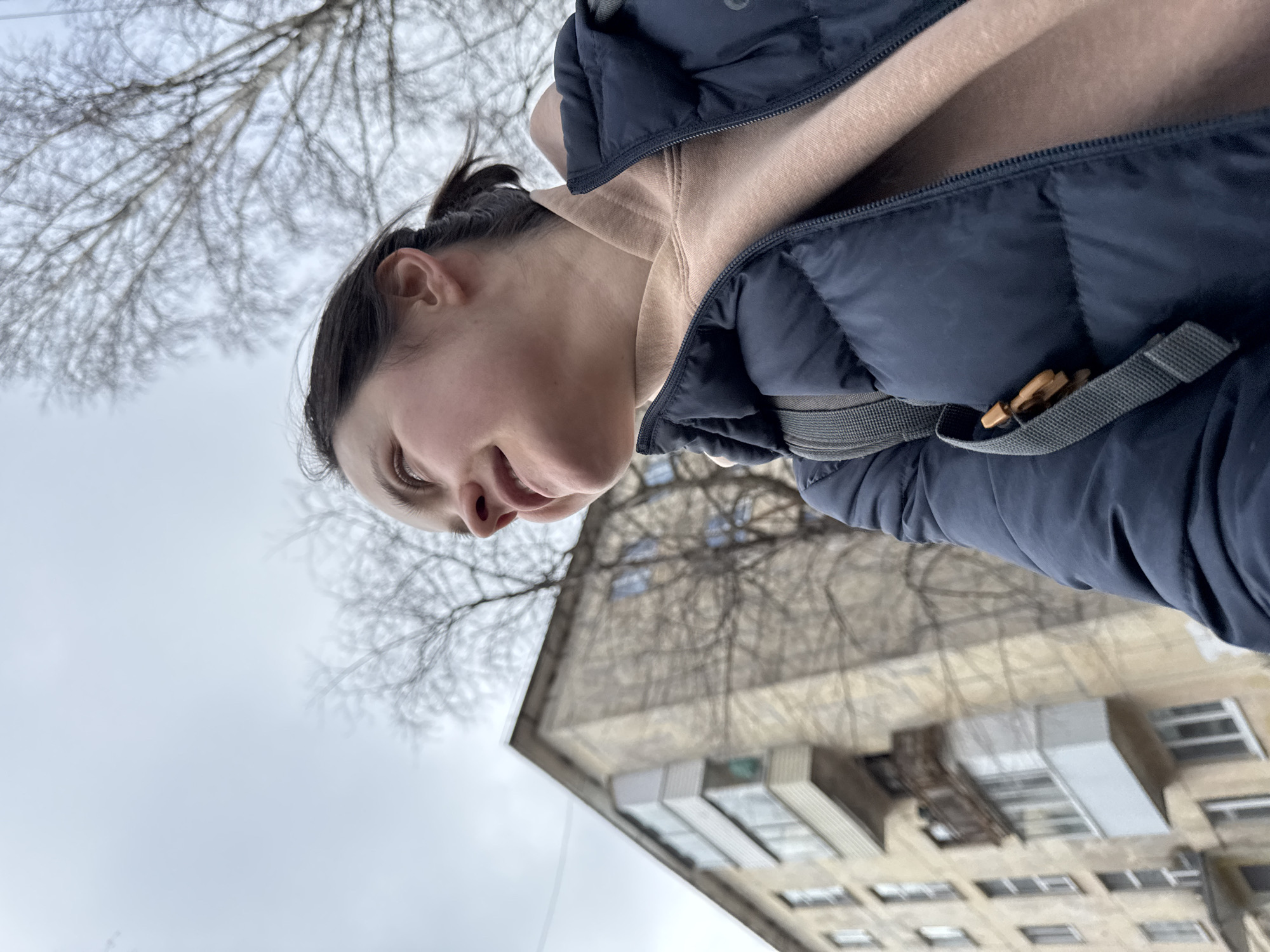 A low-angle, close-up shot depicts a person, likely female, from the upper torso to just above the eyes, looking downwards with a faint smile or smirk. Her dark hair is pulled back, revealing her ear. She is wearing a dark blue puffer jacket, unzipped to reveal a light brown or beige hooded sweatshirt underneath. A strap with a small, wooden-like toggle or fastener is visible on the right side of her jacket.
The background consists of a cloudy, overcast sky, against which the intricate, leafless branches of a tree are silhouetted, suggesting a colder season like late autumn, winter, or early spring. Behind the tree and person, a multi-story building, likely an apartment block, is visible, featuring numerous windows and what appear to be balconies or external fixtures. The architectural style hints at an urban setting, consistent with the location context of Izhevsk, Russia. The lighting is diffused, indicating an overcast daytime. There is no visible text in the image.