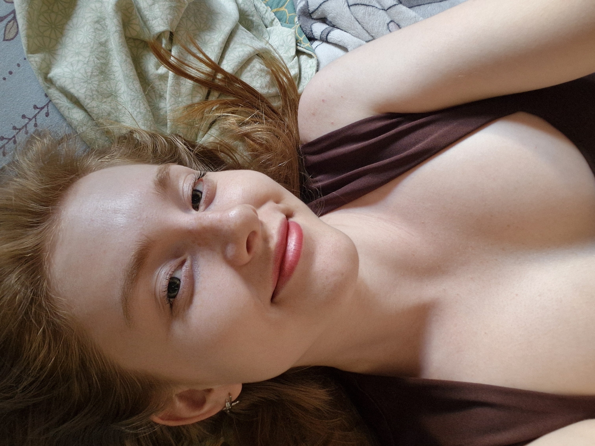 A woman with red hair lies on a bed. She has fair skin, green eyes, and red lipstick. She is wearing a brown tank top. The bed is covered with a light green and blue patterned sheet and a grey blanket. The location is not discernible from the image.