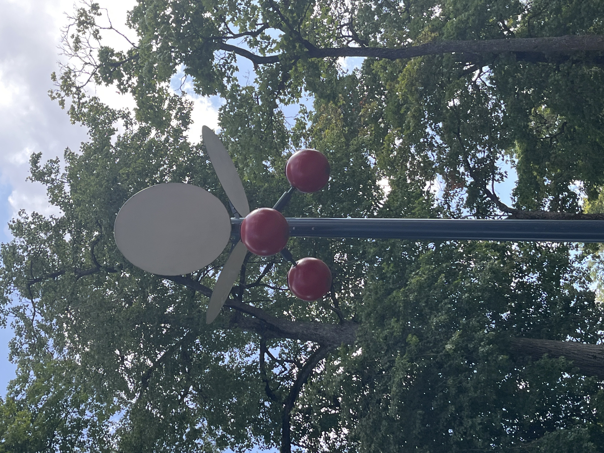 The image shows a close-up view of an outdoor sculpture against a backdrop of lush green trees and a partially cloudy sky. The sculpture is mounted on a dark pole and features a large, light-colored oval shape at the top, resembling a halo or an abstract head. Below this oval, three bright red spheres are suspended, two on the sides and one in the center, creating a playful, almost festive appearance. The sculpture is positioned in front of mature trees with dense foliage, suggesting a park or a garden setting. The lighting indicates it is daytime, with some sunlight filtering through the leaves. The location is identified as Jõhvi, Estonia. There are no people or animals visible in the image. The composition focuses on the sculpture and its natural surroundings.