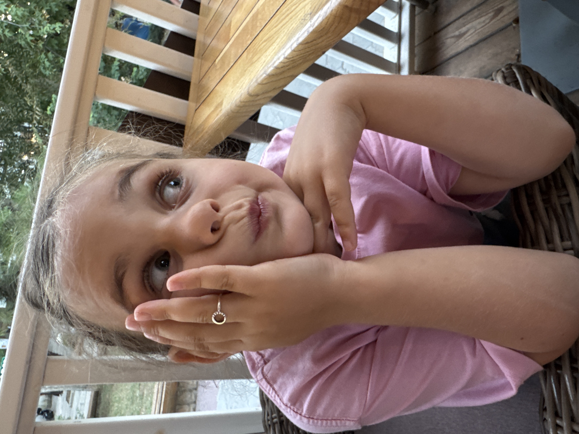 A young girl is shown from the chest up, sitting outdoors at what appears to be a table and chairs on a deck. The girl is wearing a pink t-shirt and has her hands propped up on her chin, with her left hand touching her face. She is wearing a ring on her left ring finger. Her mouth is pursed, and she is looking to the right. In the background, a wooden railing and table are visible, as well as greenery and what looks like a blue shirt worn by a person in the distance. The lighting suggests it is daytime. The location is not determinable from the image.