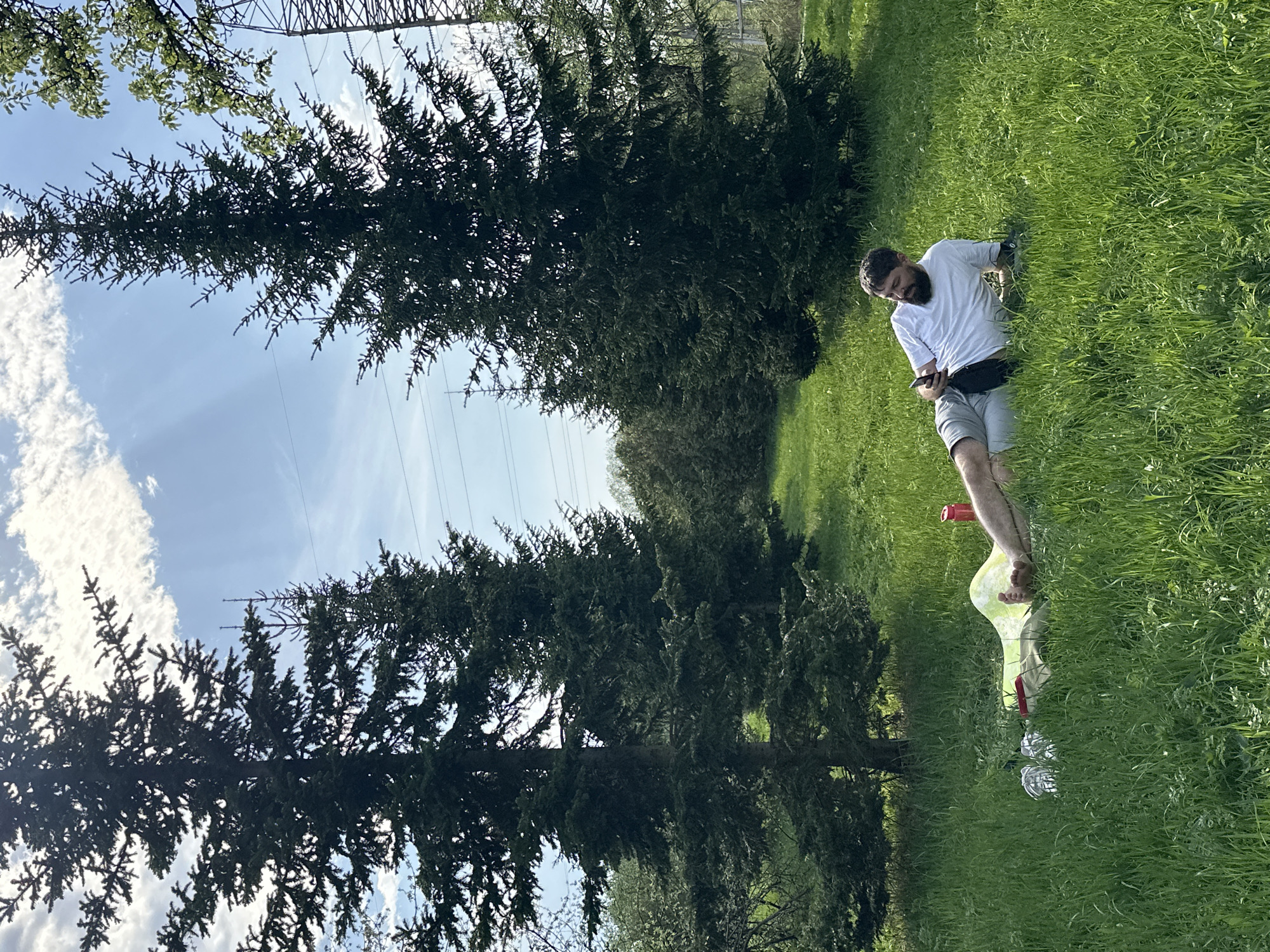 A man with a beard, wearing a white t-shirt and gray shorts with a fanny pack, is sitting in a grassy field, looking at his phone. Beside him is an empty wine bottle with a red cap and what appears to be a crumpled tablecloth. Two tall evergreen trees flank the scene, reaching towards a sky with scattered clouds and power lines. The location is Moscow, Russia.