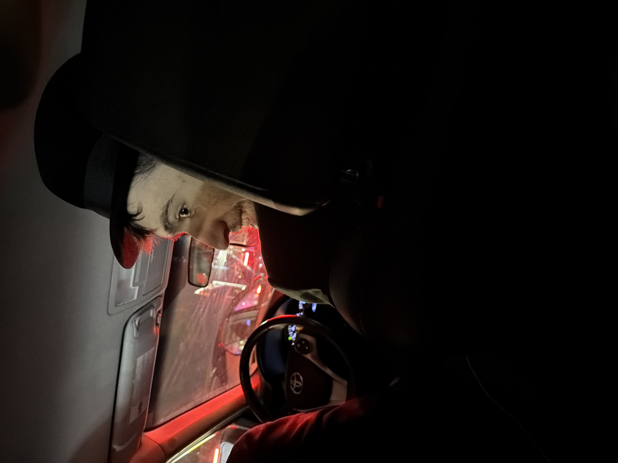 A person with light skin and a dark beard is wearing a black hat inside a car. The person is looking toward the left side of the frame. The car's interior is visible, including the steering wheel with the Toyota logo, dashboard, and window. Red and white lights are visible outside the car window. The image appears to have been taken at night. The location in the city of Fort Lee, United States of America, is not discernible from the image.
