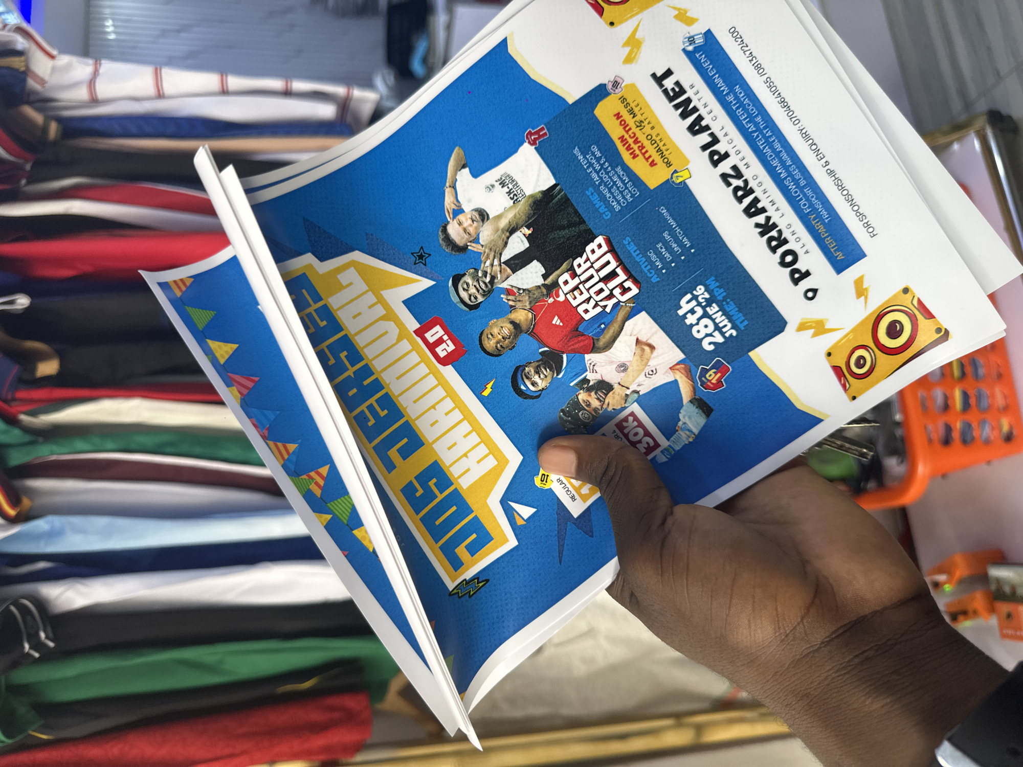 The media file is a close-up shot of a person's hand holding a flyer for an event titled "PORKARZ PLANET" which is also referred to as "JOS JERSES KARNNIVAL". The flyer is predominantly blue with yellow and white text and graphics. It features images of several people, likely football players or celebrities, in athletic attire.
The flyer promotes various activities including music, dance, match-making, link-ups, and games such as snooker, table tennis, chess, ludo, and PES. A main attraction advertised is a "RONALDO VS MESSI (FANS BATTLE)". The event is scheduled for "28th JUNE '26" at "1 PM".
Additional information on the flyer includes details about an after-party, transport arrangements, and contact numbers for sponsorship and inquiries. The event appears to be taking place in Jos, Nigeria, as indicated by "JOS" in the title and "LAMINGO MEDICAL CENTER" as a location cue. The background of the image shows stacks of folded clothing, suggesting the photo might have been taken in a retail or storage environment. The lighting suggests an indoor setting, possibly during daytime. The overall tone of the flyer is energetic and celebratory, aiming to attract attendees to a sporting and social event.