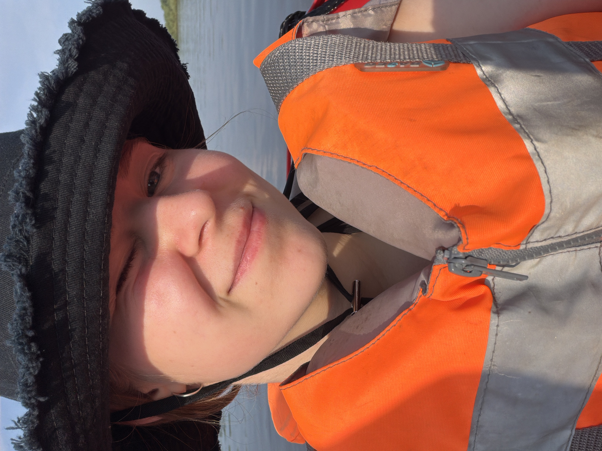 The image is a close-up selfie of a young person wearing a black bucket hat and an orange life vest. The person is looking directly at the camera with a slight smile and one eye closed, suggesting a wink or squint. The background shows a body of water with a faint shoreline in the distance. The lighting indicates it is daytime, with the sun casting a warm glow on the person's face. The scene appears to be on a boat or near the water, with the life vest suggesting recreational activity. The location is described as being in Kalino, Russia, implying an outdoor setting by a river, lake, or sea.