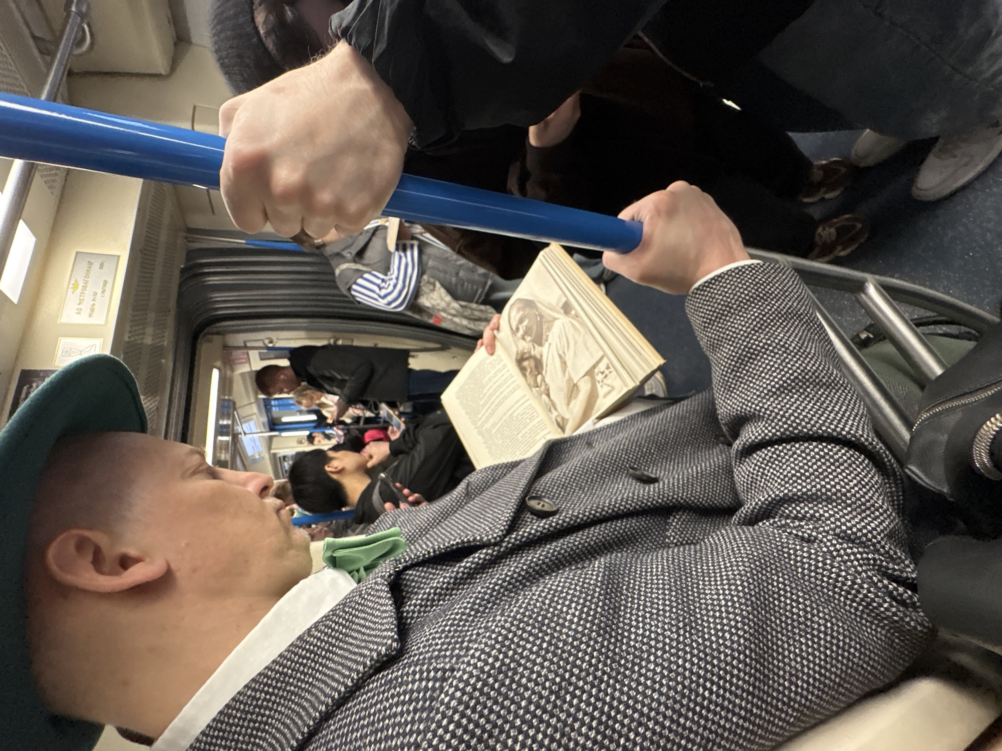 The image depicts the interior of a Moscow Metro train car in Russia. Several people are visible, including a man in a green hat and patterned blazer who is reading a book. Another person is holding onto a blue handrail. Other passengers are seen sitting or standing in the background. The train car features metal seating frames and advertising panels on the walls.