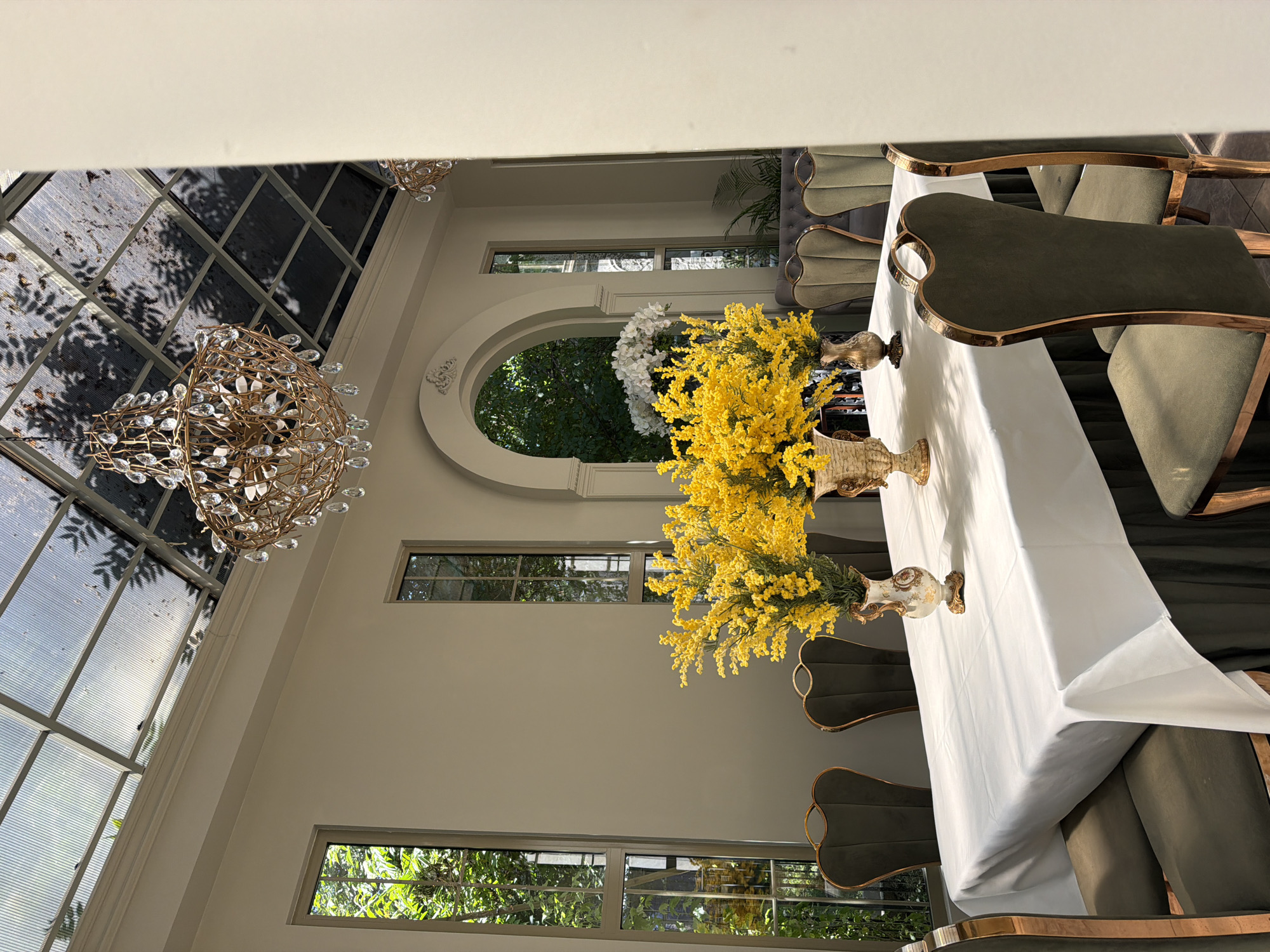 The image shows an indoor setting with a table set for a meal. The table is covered with a white tablecloth and adorned with flower arrangements featuring yellow flowers in vases. Around the table are chairs with a distinctive design. A chandelier hangs from the ceiling. Natural light filters through a large window with a patterned frame, suggesting a sunny outdoor setting. The location is Bishkek City, Kyrgyzstan.