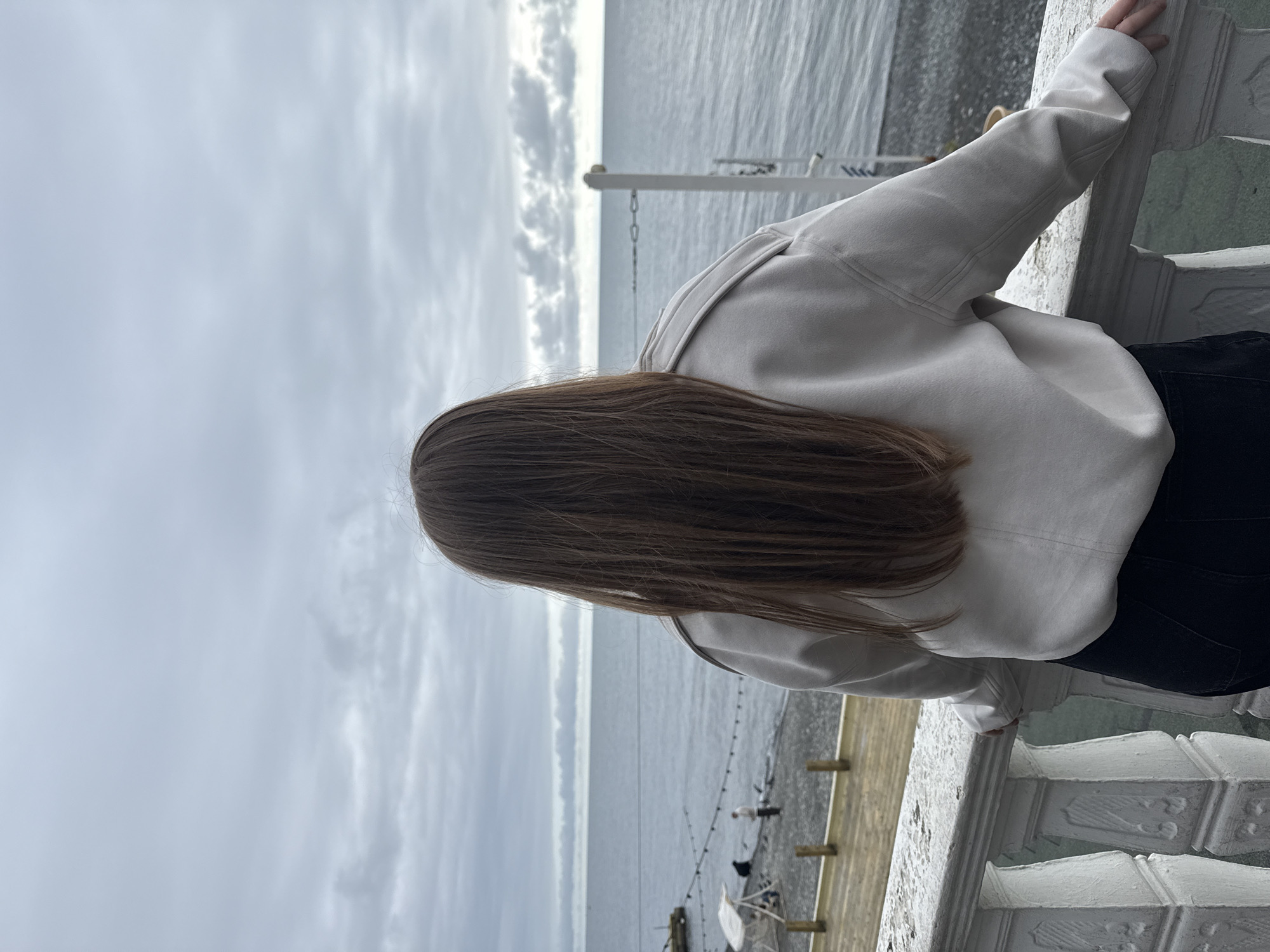 A person with long brown hair, viewed from behind, stands on a balcony or terrace overlooking the Black Sea in Sochi, Russia. The individual wears a light-colored, long-sleeved top and dark pants, with their left hand resting on a weathered white balustrade featuring ornate carvings. Below the balustrade, a wooden platform transitions to a dark pebble beach. On the beach, several indistinct figures are present, including one person in white standing upright and another crouched figure. A white canopy structure and a string of lights are also visible on the shoreline. A short wooden pier extends into the water on the left, and a white pole with chain links is partially submerged on the right. The sea is calm, with a grey-blue hue under an overcast sky filled with layered clouds. The horizon line is clearly defined.