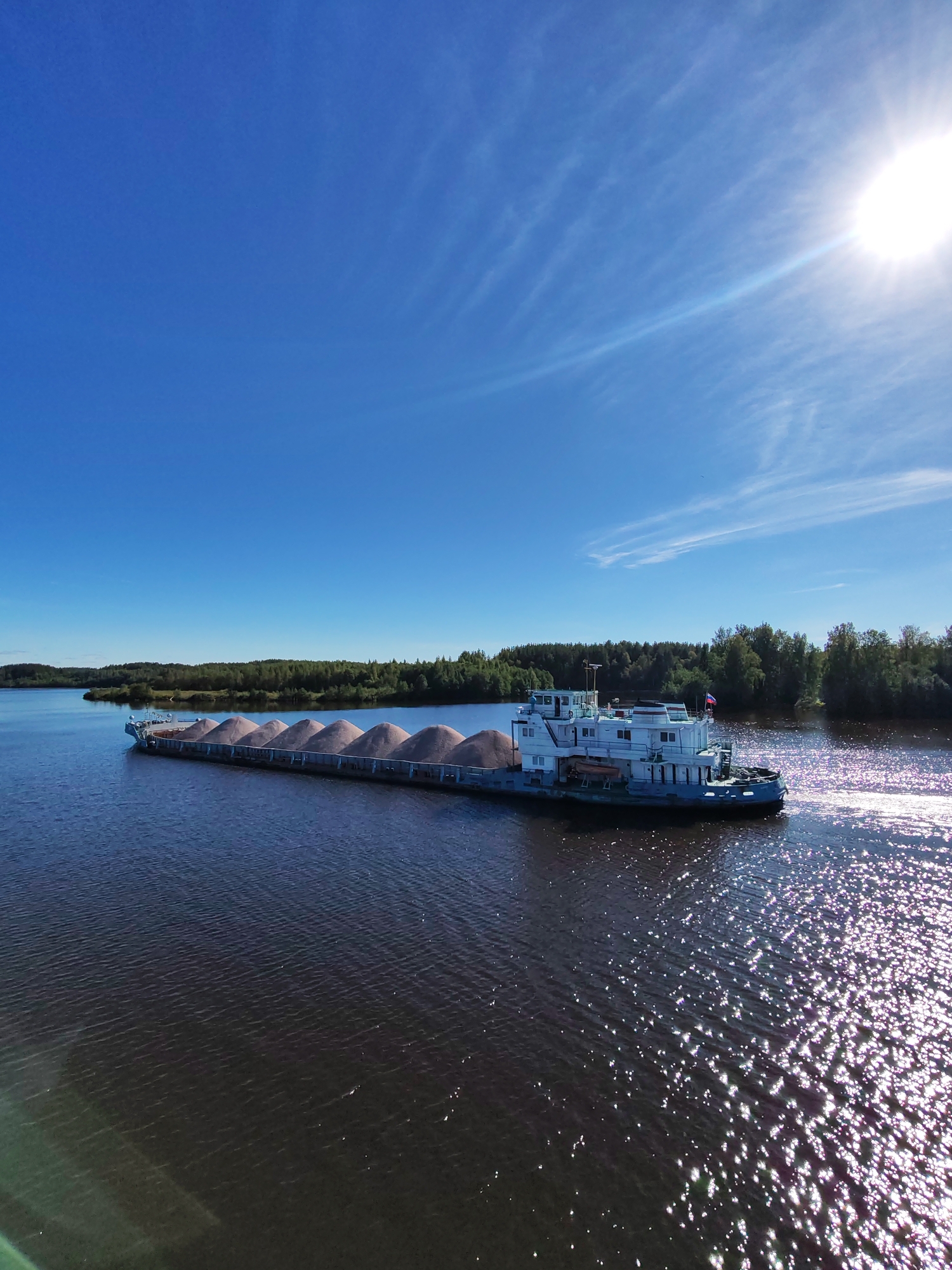 An aerial, wide-angle shot captures a bright, sunny day over a wide, dark river in Belousovo, Russia. The sky is a vibrant, cloudless blue, with a few thin, wispy clouds streaking across the upper right quadrant where the sun shines intensely. The sun's glare creates a sparkling effect on the water's surface, which reflects the light in a shimmering pattern.
Dominating the midground is a long, flat barge heavily laden with a beige, granular substance, likely sand or gravel, piled in neat, triangular mounds. The barge is being pushed by a white and blue tugboat, which is positioned at the stern of the barge. The tugboat has several decks and windows, and a Russian flag is visible on its superstructure. The barge is moving forward, creating a visible wake in the dark water.
The riverbanks are lined with dense green trees, forming a lush, natural backdrop. The trees on the left bank appear further away, forming a rolling landscape, while those on the right bank are closer and more prominent. The water appears calm, with subtle ripples. The overall impression is one of industrial transport on a beautiful, clear day.