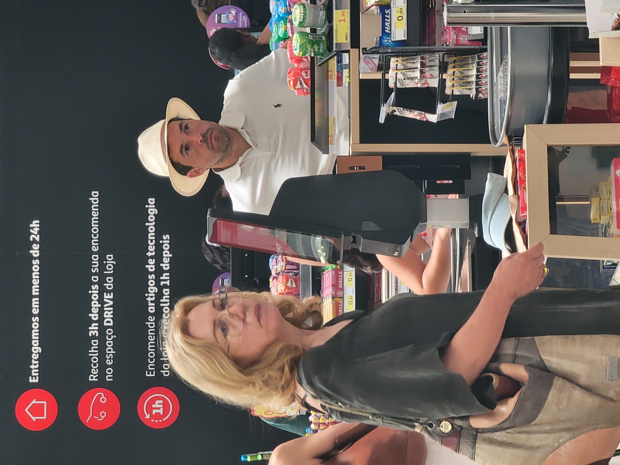 This image was taken inside a retail store, likely a supermarket, in Faro, Portugal. In the foreground, a woman with blonde, shoulder-length hair and glasses is seen from the chest up, wearing a dark green V-neck top and carrying a large, tan and brown handbag. She is positioned on the left side of the frame, facing to the right. Behind her and to her right, a man wearing a light-colored fedora and a white polo shirt with a dark logo is visible from the chest up, looking off to the right. He appears to be an employee or another customer.
The background reveals shelves stocked with various products, including candy, gum, and possibly pharmaceuticals, indicated by packaging for "Halls" and other brands. A checkout counter or display stand is also present, with a black monitor and some product displays. Notably, a black graphic on the wall behind the individuals features icons and text in Portuguese, reading "Entregamos em menos de 24h" (We deliver in less than 24 hours), "Recolha 3h depois a sua encomenda no espaço DRIVE da loja" (Pick up 3 hours after your order at the store's DRIVE area), and "Encomende artigos de tecnologia da loja e recolha 1h depois" (Order technology items from the store and pick up 1 hour later). This suggests the store offers various convenient services. The overall lighting is bright, characteristic of an indoor retail environment.
