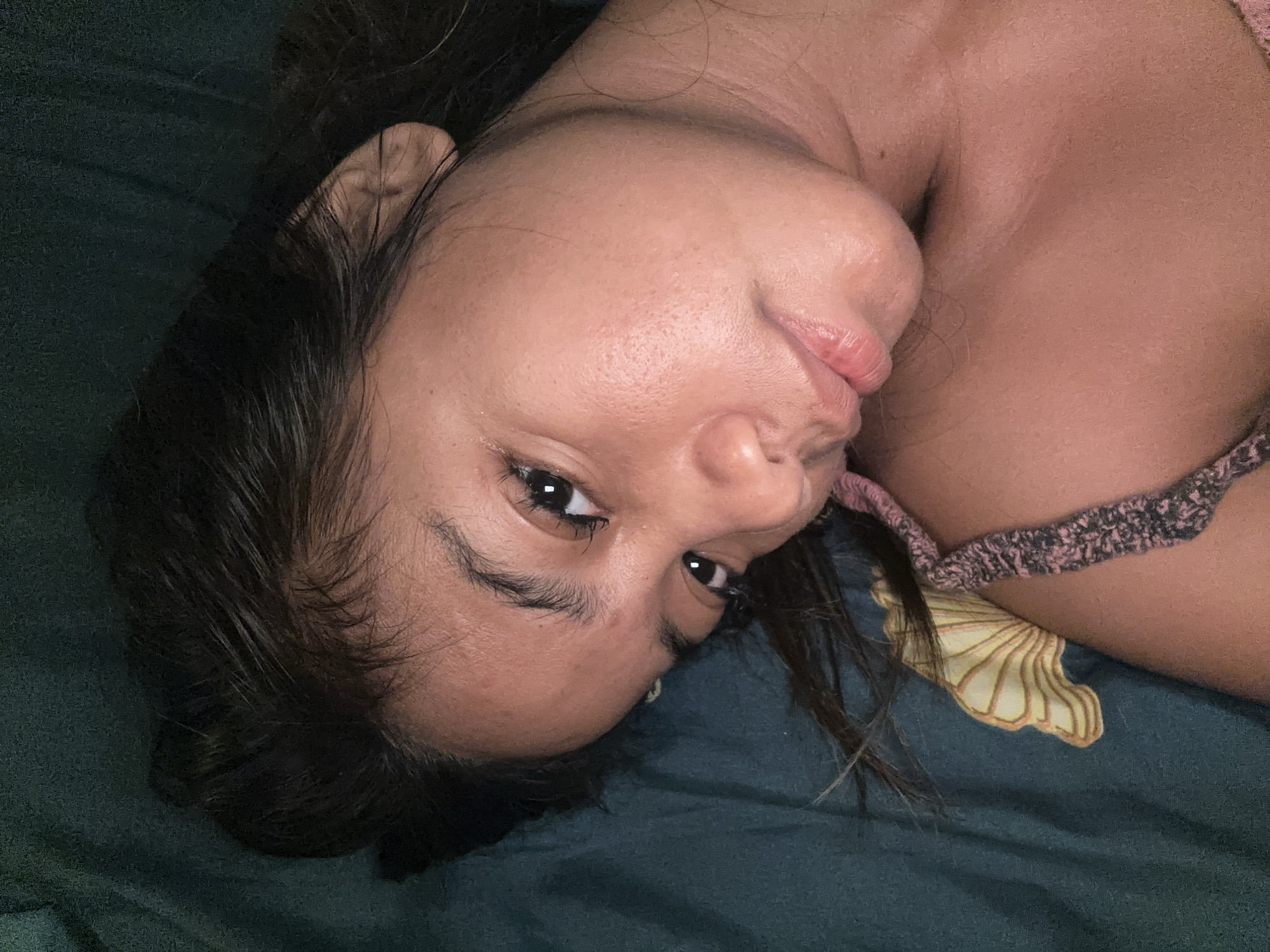 This is a close-up, vertically oriented, low-angle selfie of a young woman lying down, with her head tilted. She has long, dark hair that is spread out around her face and shoulders. Her skin is a warm, tan tone, and she appears to be wearing minimal makeup. Her eyes are dark and expressive, and her lips are slightly parted. She is wearing a pink and gray textured garment, possibly a top or bra, with a scalloped edge visible on the right side of the frame. The background consists of a dark green fabric that appears to be a bedsheet, with a subtle pattern of light yellow, shell-like shapes. The lighting suggests an indoor setting, possibly in the evening or in a dimly lit room. The woman's expression is relaxed and perhaps a little drowsy, giving a sense of intimacy and informality. There is no visible text in the image. The location context is Sukawati, Indonesia.
