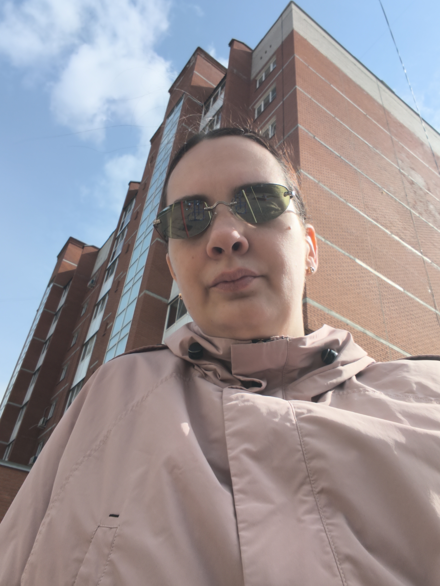 The image shows a person wearing sunglasses and a light pink jacket, with a tall brick building and a blue sky in the background. The person is positioned in the foreground, and the camera is angled upwards towards the building. The building appears to be a multi-story residential structure with windows and a light-colored top section. The scene is outdoors, under natural daylight.
