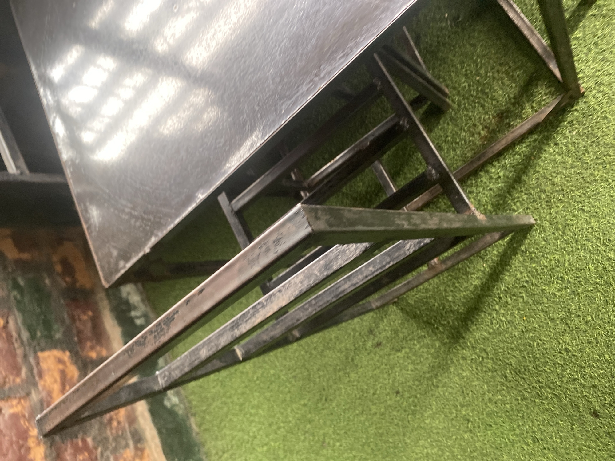 The image displays a metallic table with a dark, textured surface, reflecting light in wavy patterns. The table is supported by a dark, angular metal frame, part of which is angled upwards. The table and its frame are situated on a bright green, artificial turf surface, suggesting an outdoor or recreational setting. To the left of the table, a brick wall with visible texture and color variations is present, indicating a bordering structure or boundary.
The overall scene appears to be a still life of a table and its surroundings, with no people or active events depicted. The lighting suggests it might be daytime, given the bright reflections on the table, but the specific time is not discernible. The location cues of the artificial turf and brick wall don't provide definitive information about the exact setting beyond what's visible. There is no visible text in the image. The image is likely taken in Jalingo, Nigeria, as per the provided location context. The composition is somewhat abstract, focusing on the forms and textures of the objects and the interplay of light and shadow.