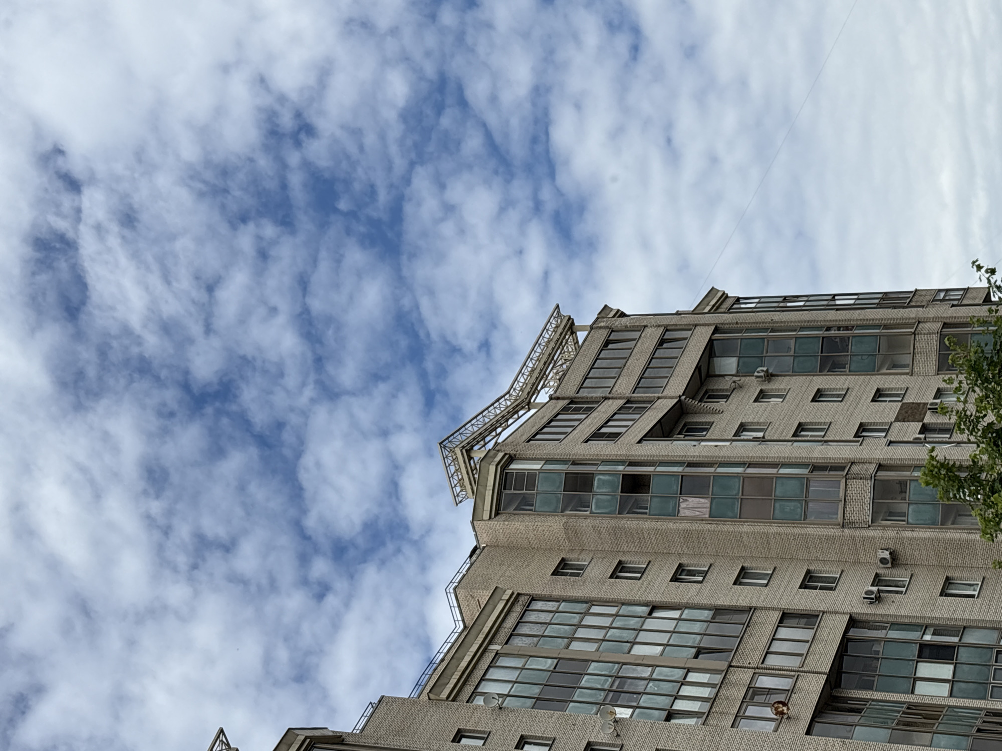 An upward-angled, medium shot captures the exterior of a multi-story brick building against a cloudy blue sky. The building, with its light beige brick facade, features numerous windows, some large and panoramic with greenish-tinted glass, others smaller and rectangular. A decorative metal structure, resembling a geometric trellis, crowns the top of the building on the right side. Air conditioning units are visible mounted on the brickwork. A thin wire stretches diagonally across the sky from the upper left to the middle right of the frame. The lower edge of the image is partially obscured by green foliage. There are no people or animals visible in the scene. The architecture suggests an urban residential or office building.