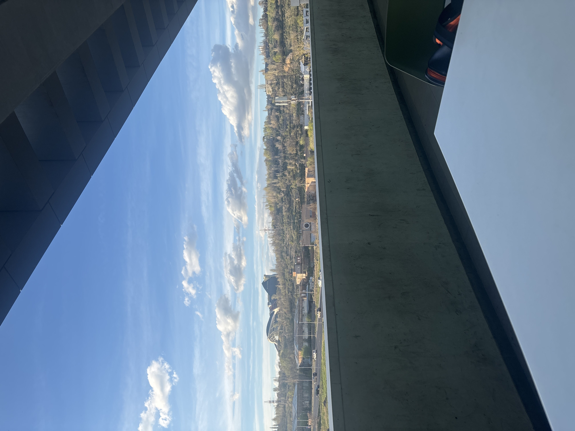 Here is a description of the image:
The image, taken from an elevated position, shows a cityscape framed by architectural elements. On the left is a structure, possibly a bridge, with a repeating pattern of horizontal supports. On the right is a concrete wall with a white surface, partially obscuring the scene. In the distance, the city of Yerevan, Armenia, stretches out with a mix of buildings, trees, and roads. The sky is blue with scattered white clouds. The Vahakn Hayots Akhparutyun (Armenian: Վահագնի Հայոց Ախպերություն) neighborhood is in the center of the image.