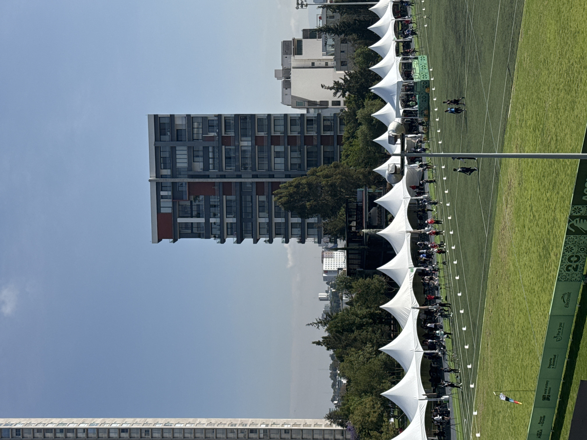 Here is a forensic description of the image:
The image shows an outdoor archery competition taking place in Puebla, Mexico. A row of white tents line the archery range. Several people are seated under the tents, presumably archers and spectators. Two people are walking on the green field toward the targets. The side of the field has green signage displaying sponsors and information about the event, including "PUEBLA," "World Archery America," "Deporte y Juventud," "Por la Paz," and "Electrolit." Buildings, including a tall high-rise, are in the background. The sky is clear and blue. The environment appears to be a city park or open area.