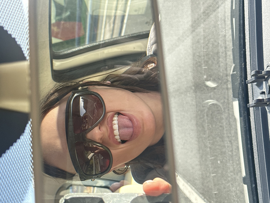 The image displays a reflection captured in a vehicle's rearview mirror. The central subject is an individual, likely female, with dark hair, visible from the forehead to the chin. She is wearing dark, wide-frame sunglasses that reflect a multi-lane road, a crash barrier, and distant foliage, indicative of a motorway or major road environment. Her mouth is open, with her tongue extended, revealing white teeth. She appears to be wearing a dark hooded garment. To the left, a blurred, reddish-orange object, possibly a hand or garment, is partially visible in the reflection. The lower portion of the mirror reflects the dusty dashboard of the vehicle and a black mounting bracket. The upper part of the mirror reflects the vehicle's headliner and the sky. The specific location within the Borough of Swale, United Kingdom, cannot be determined from the visual information provided.