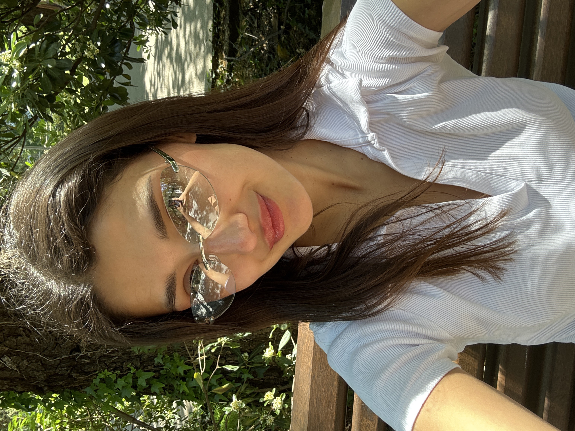 A young woman with long brown hair, wearing clear-framed glasses and a white ribbed top, sits on a wooden bench in an outdoor setting, likely a garden or park. The location seems to be the city of Bar, Montenegro as the reflection in her glasses showcases trees which are reminiscent of the location. The light suggests it is daytime. She is positioned at an angle, perhaps taking a selfie. The background includes greenery and tree bark.