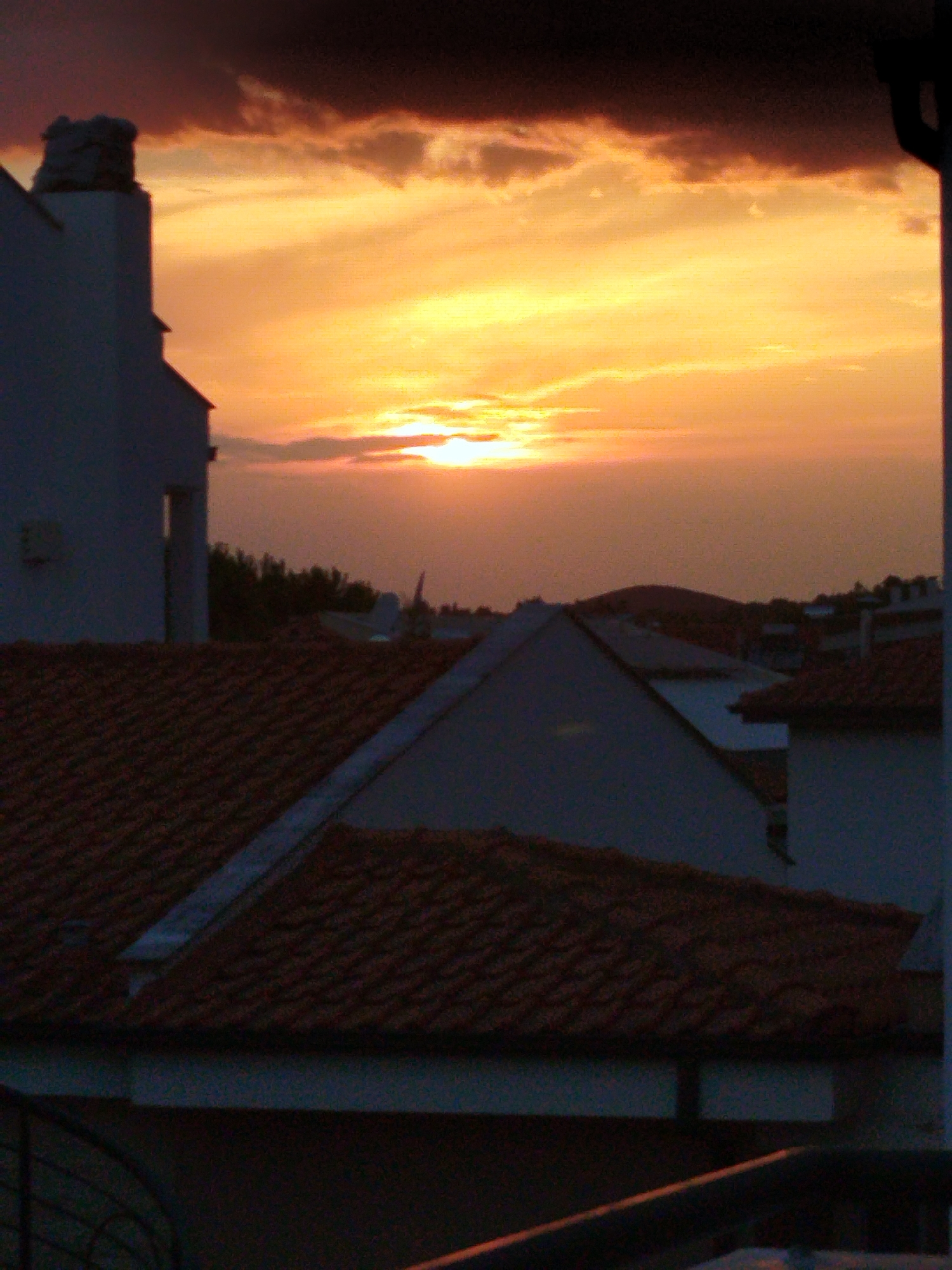 This is a vertical, medium shot of a sunset over a coastal area, likely in the Sithonia Municipal Unit, Greece. In the foreground, the dark, textured surface of a tiled roof dominates the lower portion of the image. Above the roofline, several buildings are visible. To the left, a white building with a flat roof and a visible chimney is silhouetted against the sky. Further back, a cluster of buildings with tiled roofs creates a darker, more shadowed landscape. The sky is filled with dramatic sunset colors, transitioning from deep orange and yellow near the horizon to darker shades of red and purple at the top. Wispy clouds are scattered across the sky, illuminated by the setting sun. The overall impression is one of a quiet, end-of-day scene in a residential area.