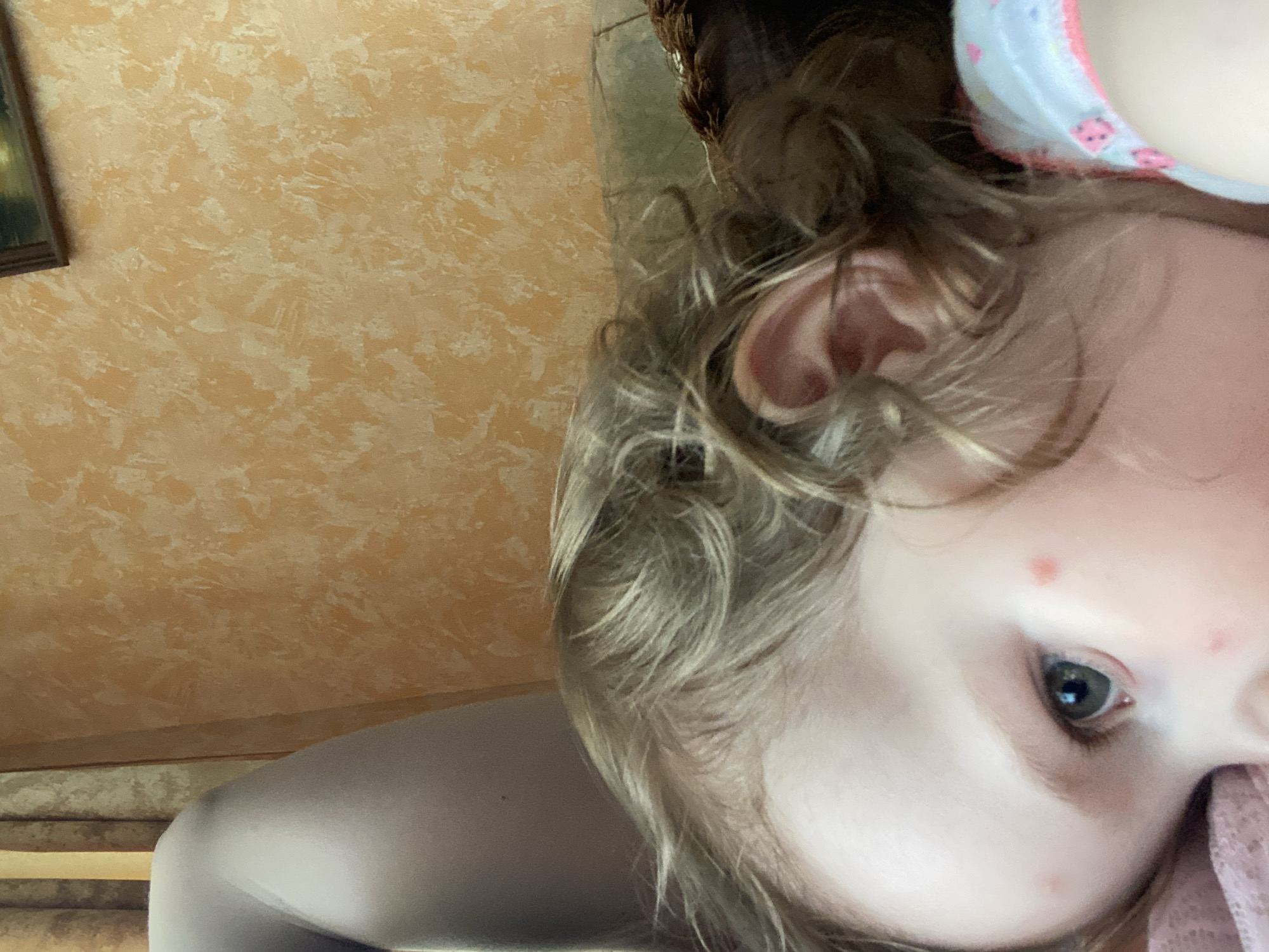 The image shows a close-up, upside-down view of a young child's face. The child has blonde hair and light-colored eyes. There are a few small red spots on the child's forehead and cheek, which appear to be pimples or minor irritations. The child is wearing a light-colored garment with pink trim and a pattern of what look like ladybugs. A pink fabric, possibly a blanket or clothing, is visible near the child's chin.
The background is a textured wall, painted in shades of orange and cream, with a framed picture visible on the left side. The lighting suggests it is daytime, and the indoor setting is warm and cozy. The child's expression is not clearly discernible due to the close-up crop and orientation. The overall impression is of an intimate, candid moment with a child.