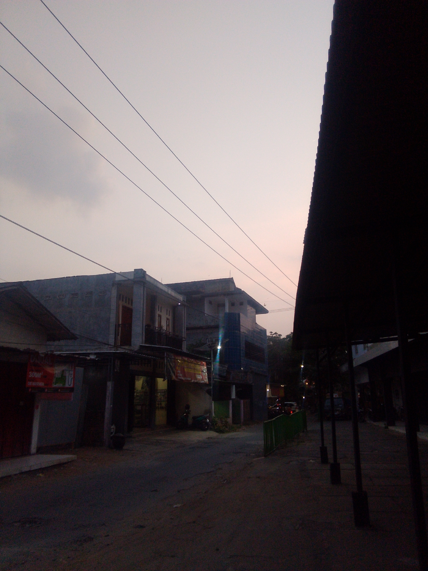 The image depicts a street scene at dusk in Trenggalek, Indonesia. The sky is overcast with a pale orange hue. Multiple power lines stretch across the sky. On the left side of the image, there are buildings with several stories. The foreground on the left shows the side of a building with a red awning. Further down the street, there are more buildings with shops. A motorbike is parked on the right side of the road. On the right side of the image, there is a dark, sloped roof structure with vertical poles supporting it. A green fence runs along the side of the street. The street surface appears to be asphalt and dirt. The scene is dimly lit, suggesting it is either early morning or late evening.