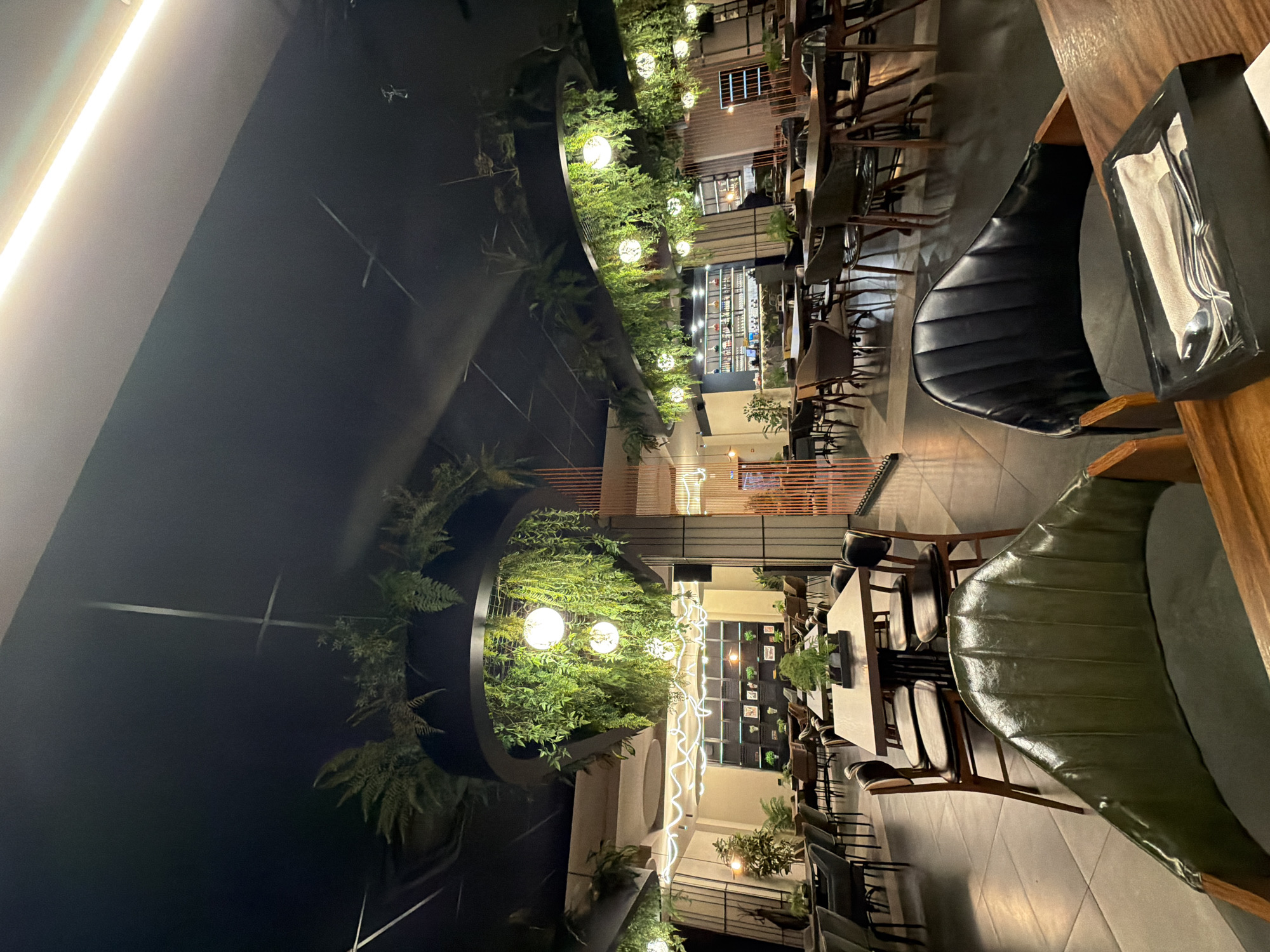 The photograph shows the interior of a restaurant in Izhevsk, Russia. The space is decorated with plants and illuminated by decorative lighting. The seating arrangement is visible with tables and chairs set up. The focus of the image is on the dining furniture, particularly the chairs with dark leather upholstery. In the background, there is a view towards a bar area, which is partially visible. The scene appears to be indoors, with a modern and stylish atmosphere. No people are visible in the image.